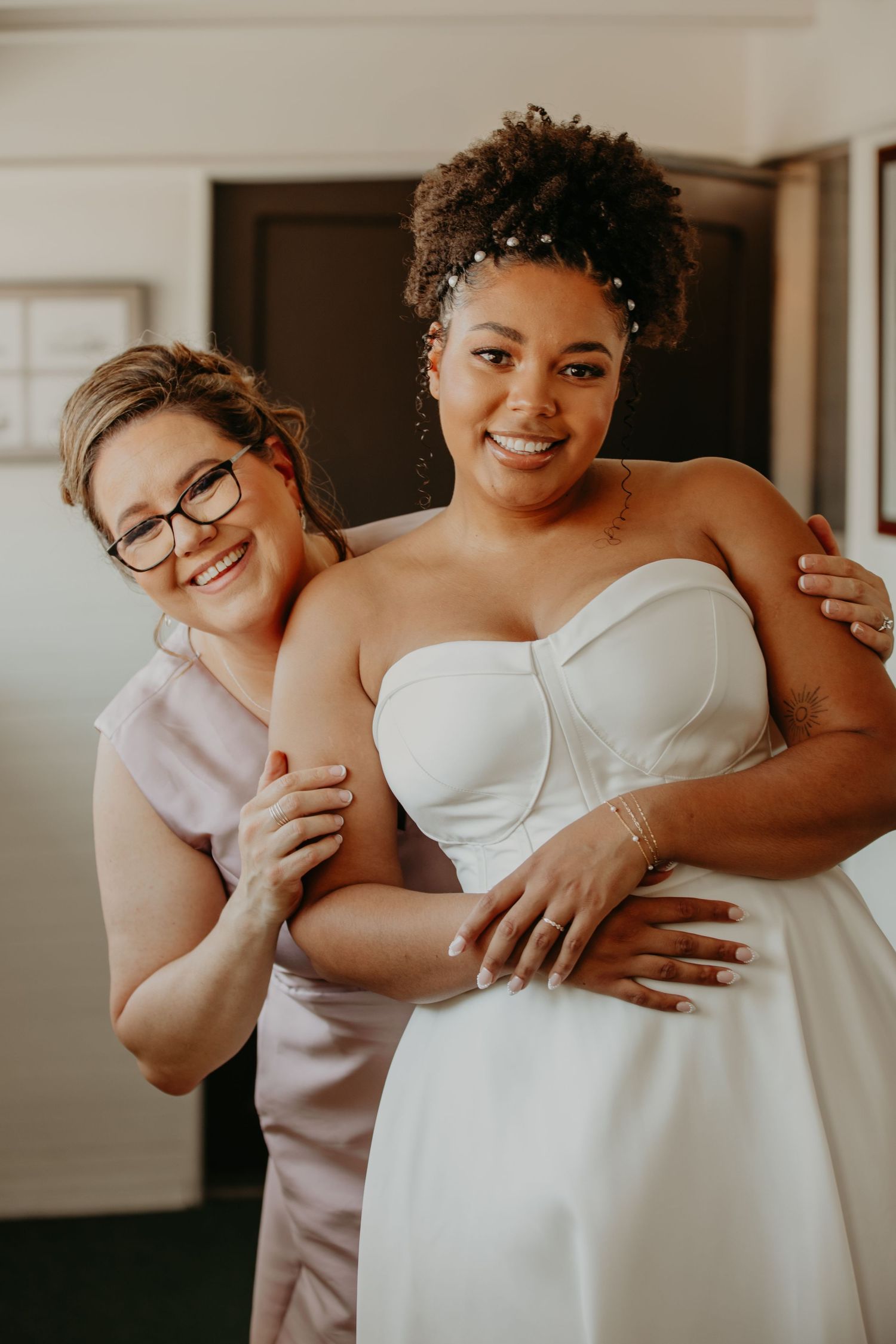 A tender getting ready moment between two people, one in a white wedding dress and one in pink.