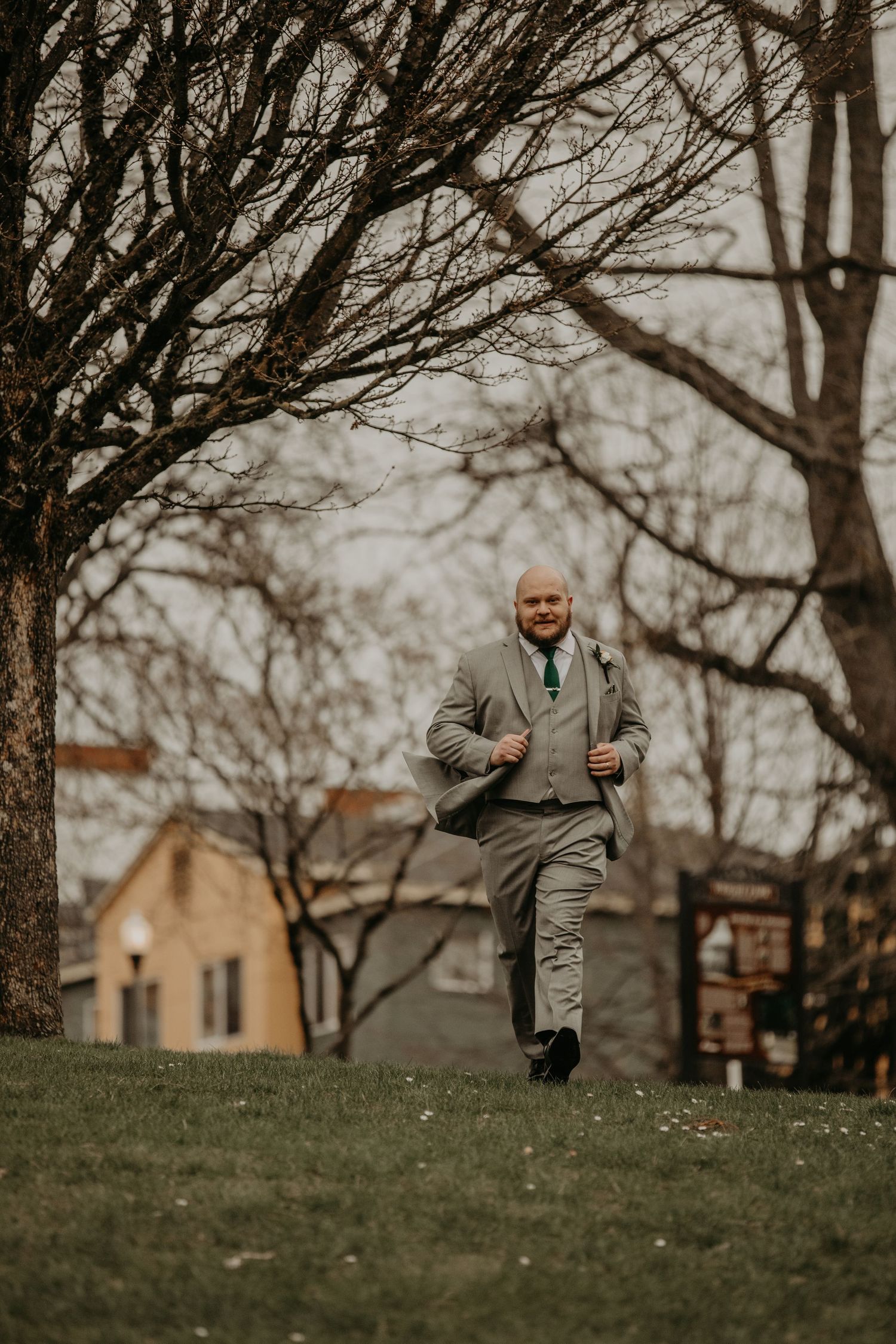 A well-dressed person in a gray suit walking on a grassy hill with bare trees and buildings in the background.