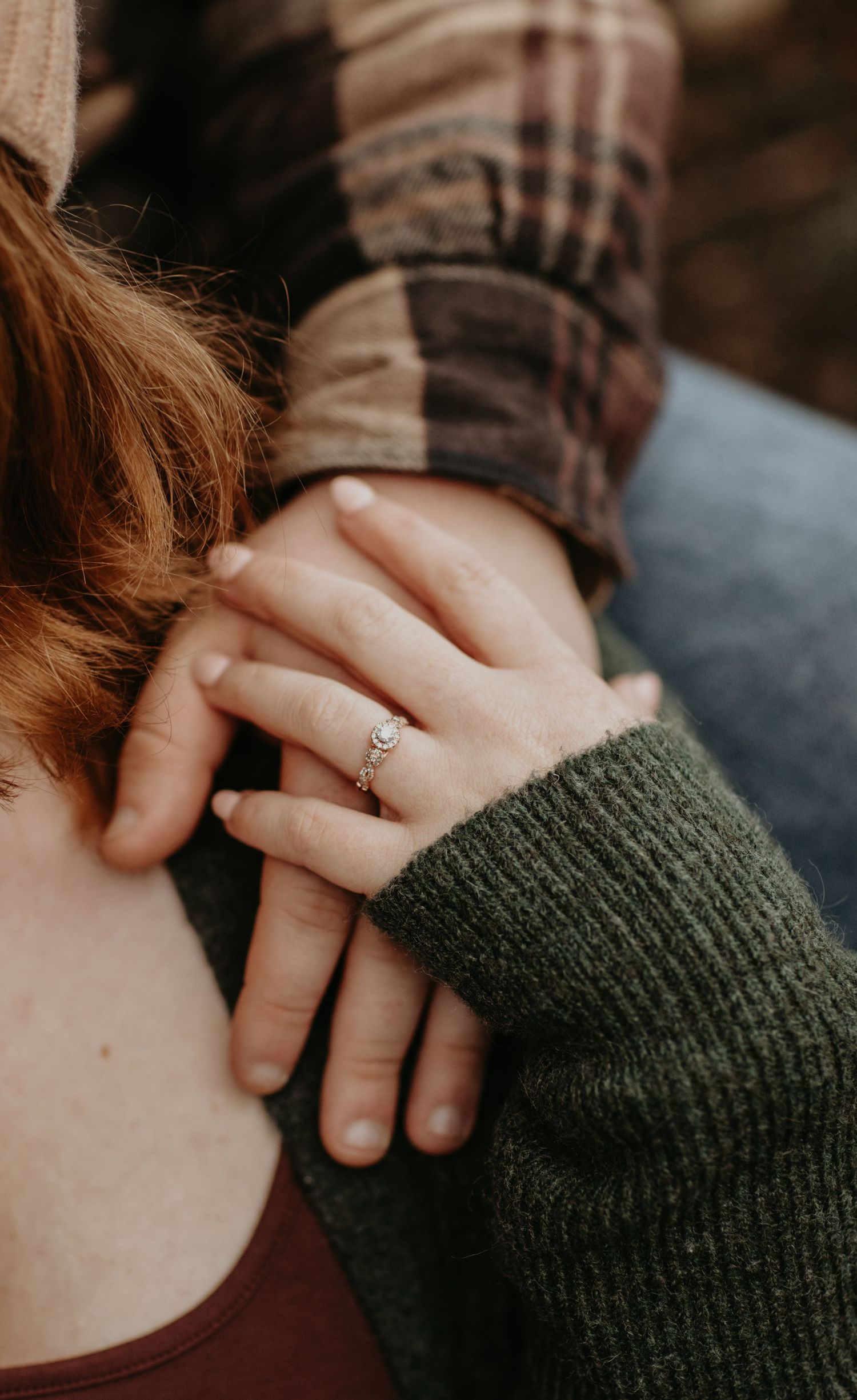 A close-up shot of hands with an engagement ring, wearing a green sweater sleeve against a plaid background.