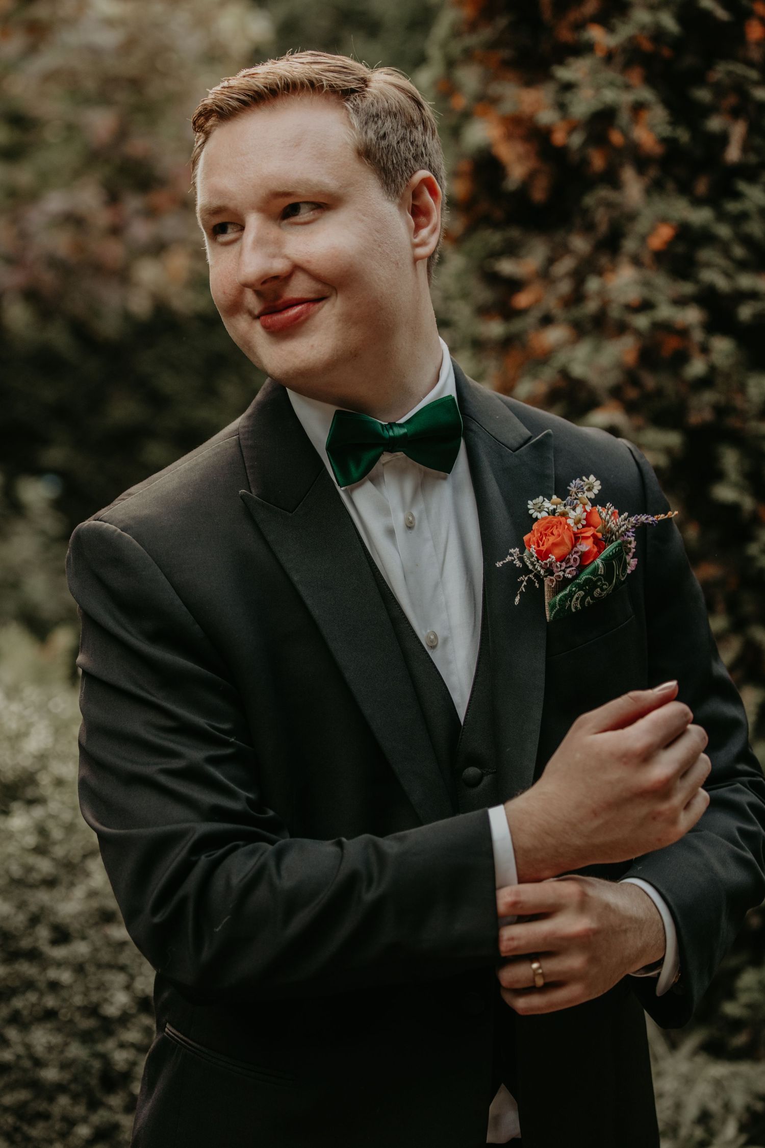 A groom in formal black tuxedo and bow tie with red boutonniere poses for a wedding photo outdoors.