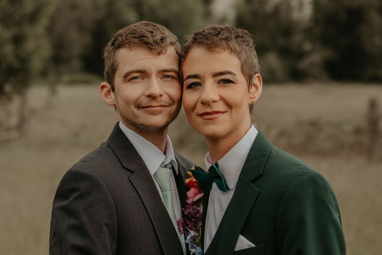 A happy couple wearing suits pose together for a wedding photo in warm golden light outdoors.