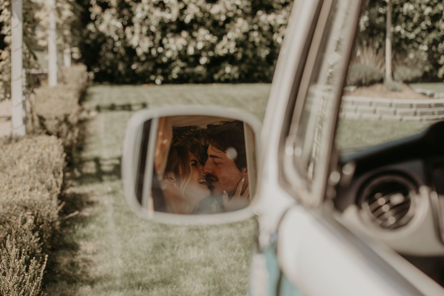 Reflection in car side mirror shows couple in a romantic moment against blurred green garden background.