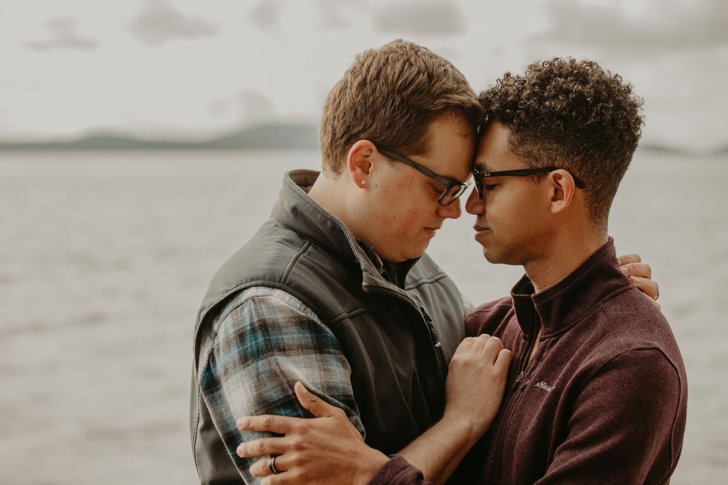 A tender intimate moment between a couple in casual wear embracing by a lake on an overcast day.