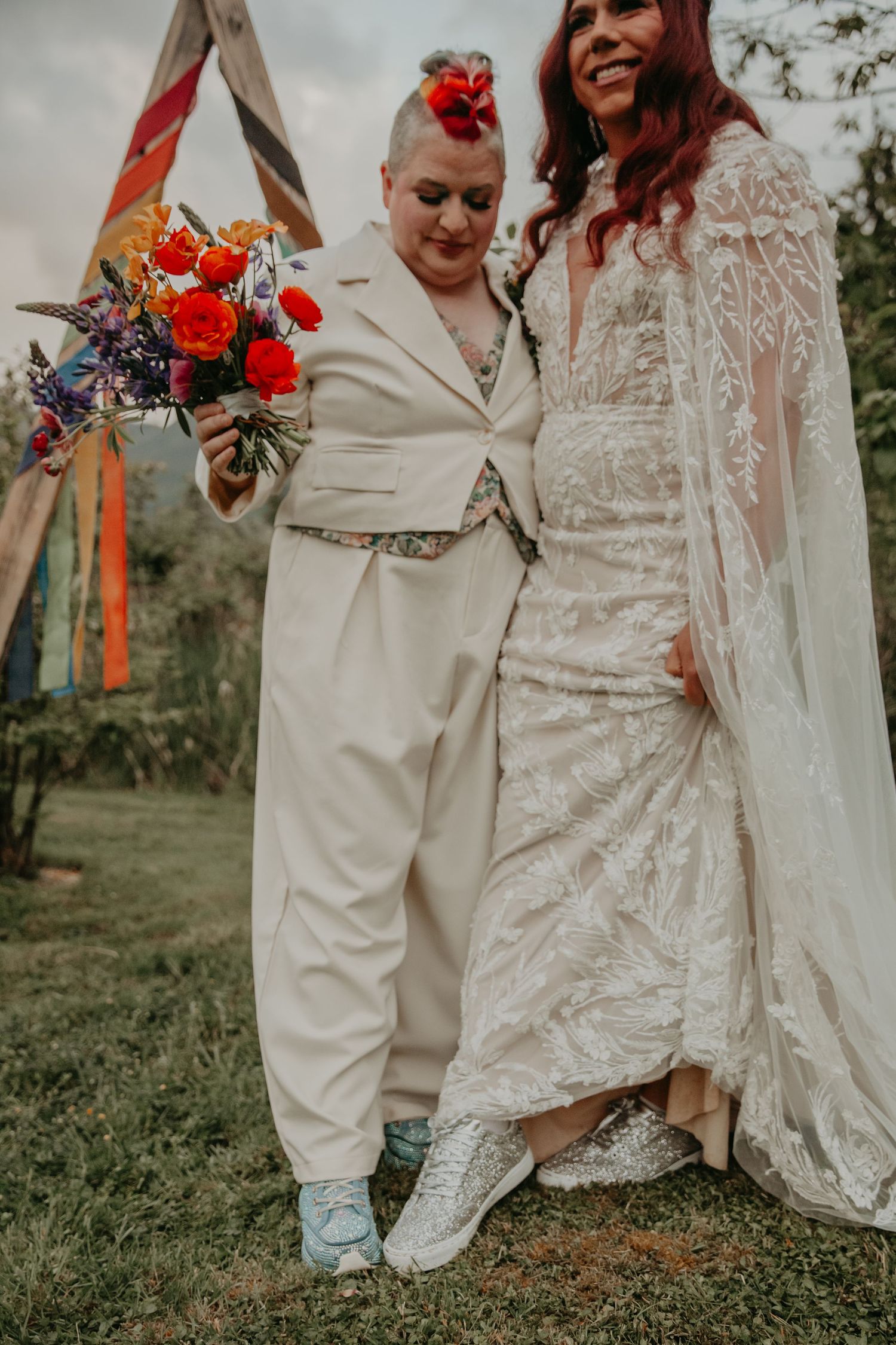 Two brides in modern wedding attire - one in a white suit and one in a lace dress - stand together with colorful bouquet.