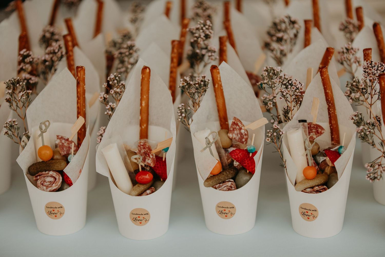 White paper cups filled with hot cocoa mix, cinnamon sticks, chocolate treats and dried flowers arranged in neat rows.