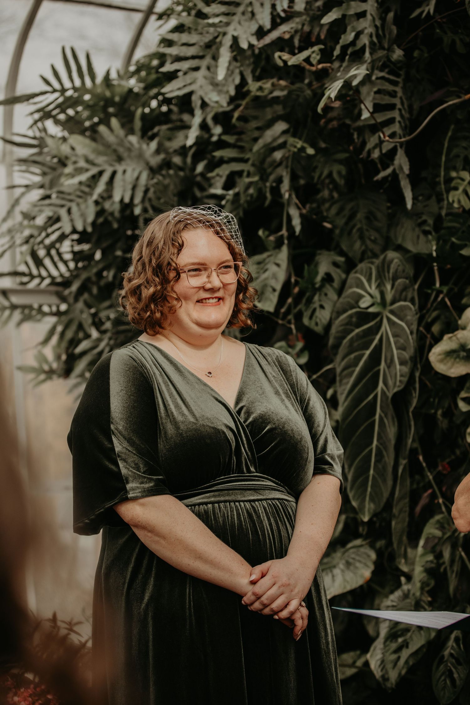 A person in a dark green velvet dress poses in front of lush tropical plants in an indoor setting.