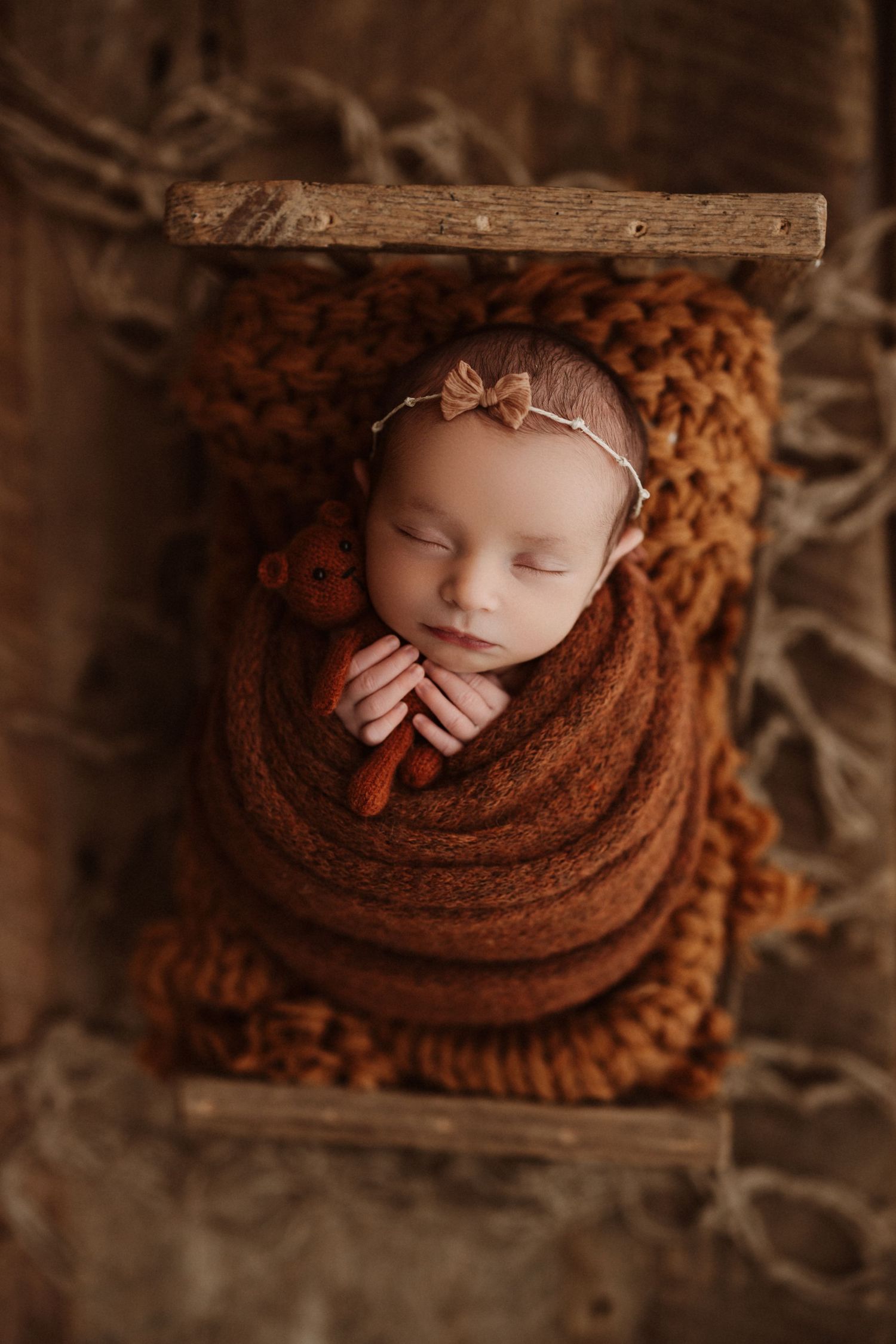 A sleeping newborn wrapped in cozy brown knit fabric rests in a rustic wooden basket.