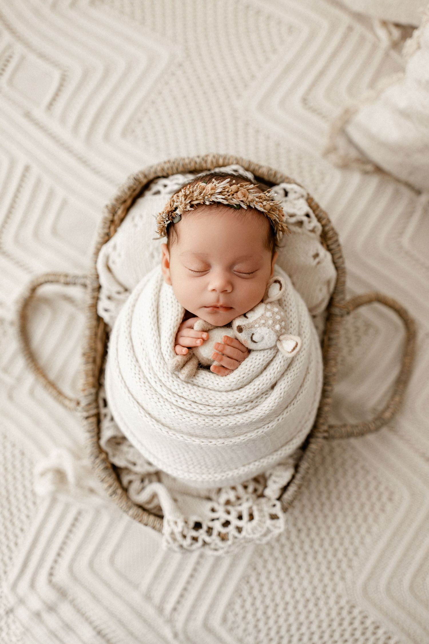 Newborn baby swaddled in white fabric sleeping peacefully in a woven basket with lace trim and floral crown.