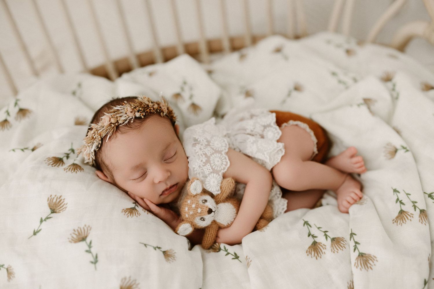 Sleeping newborn rests peacefully on white floral patterned bedding while holding a small stuffed toy.
