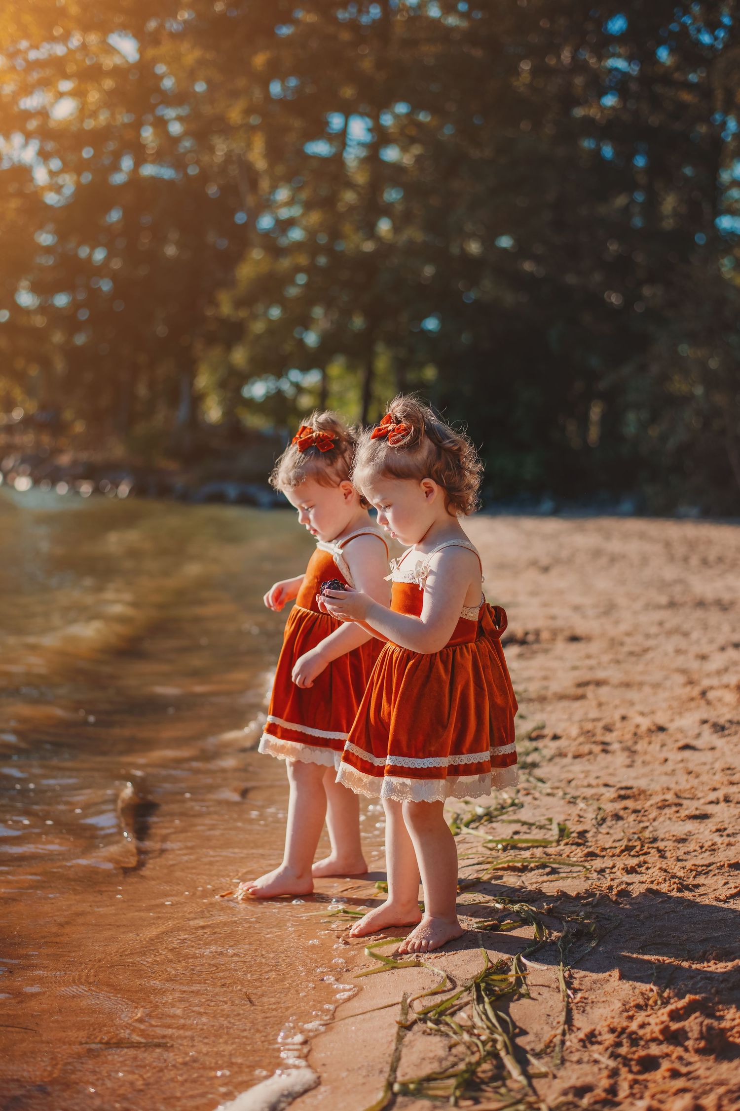 Two toddlers in matching red dresses play by riverside at sunset.