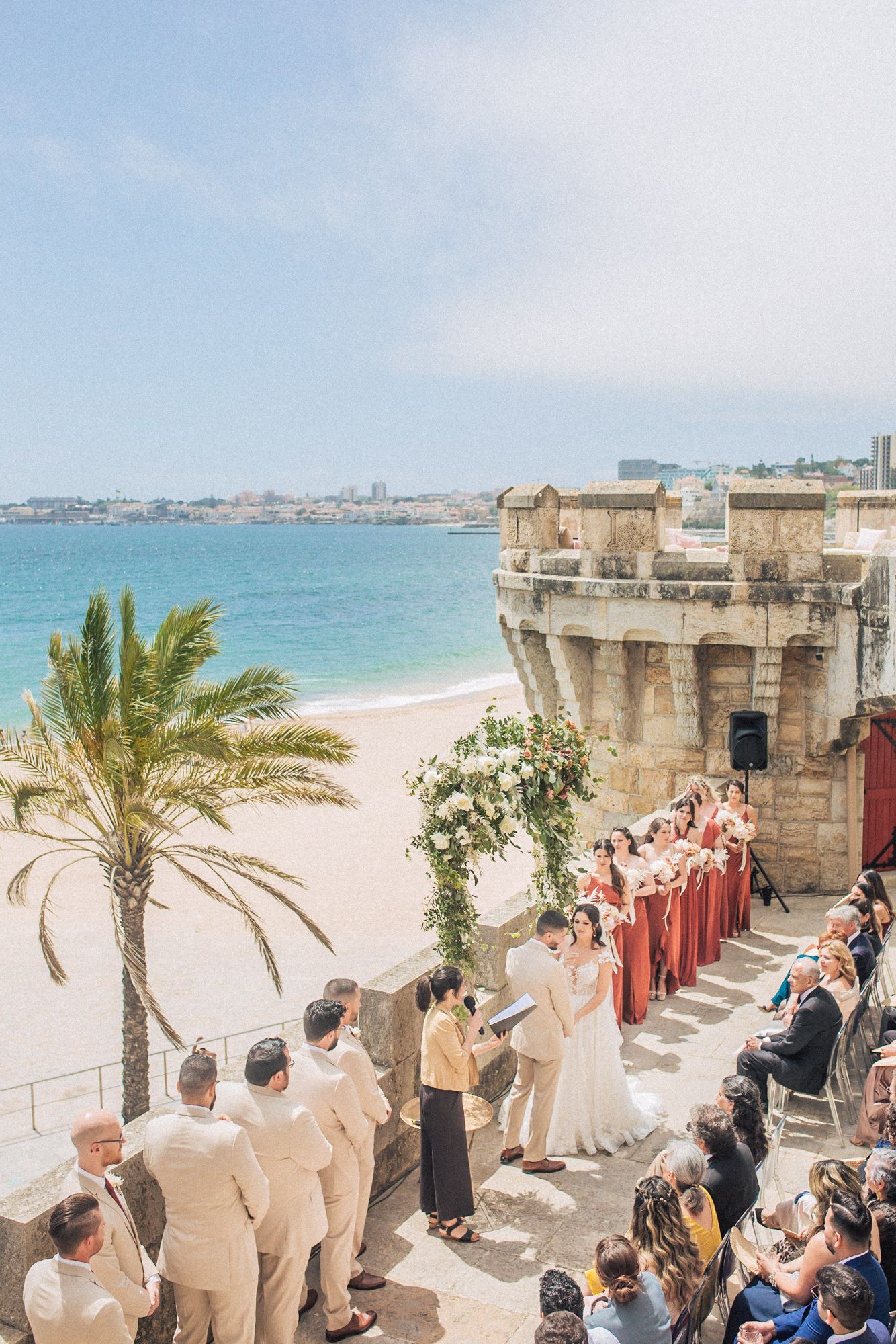 Beachfront wedding ceremony at forte da cruz with bridesmaids in coral dresses. captured by a destination wedding photographer in Portugal