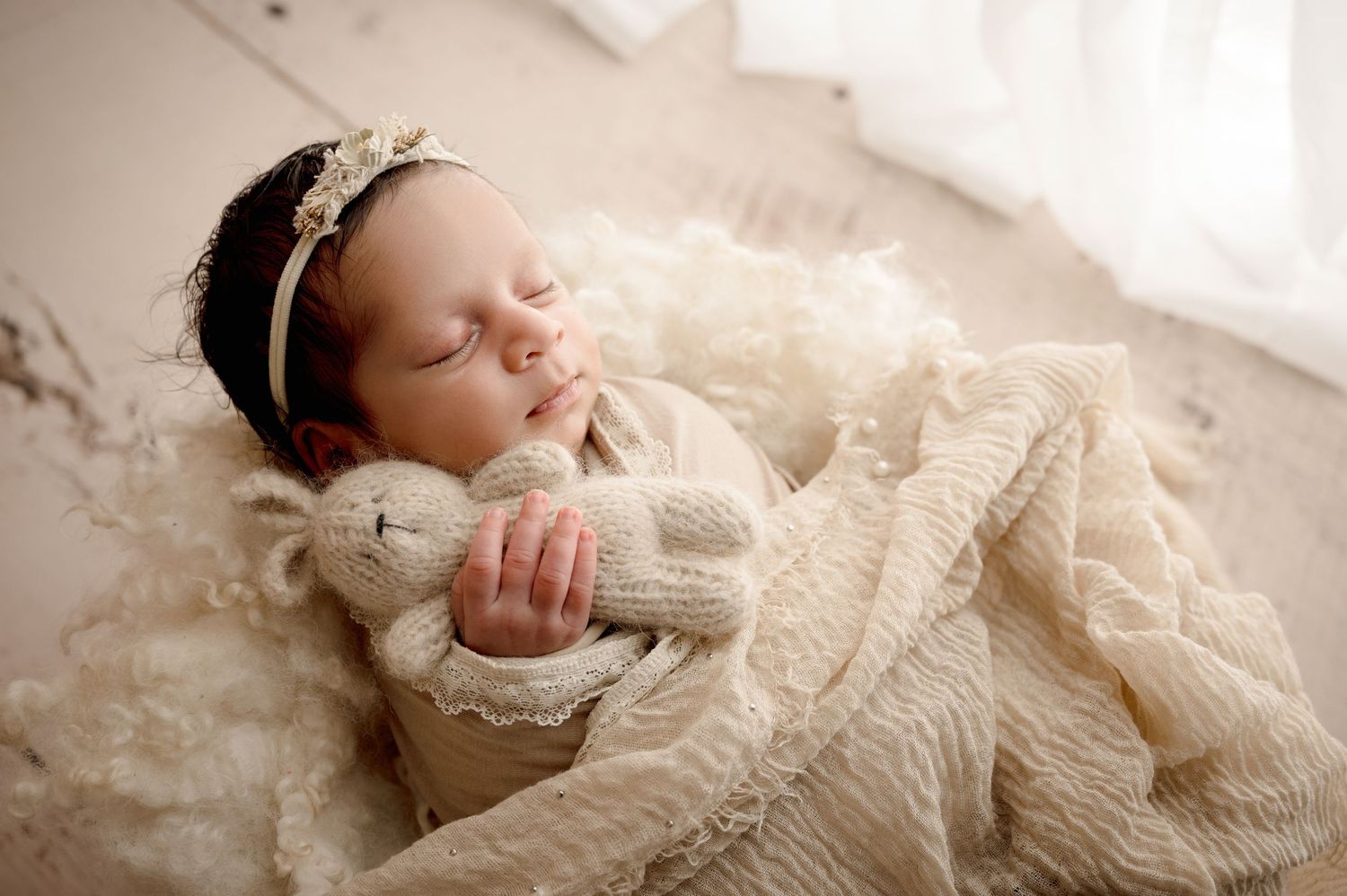 Sleeping newborn wrapped in cream knitted blanket and wearing lace headband rests on soft textured white fabric.