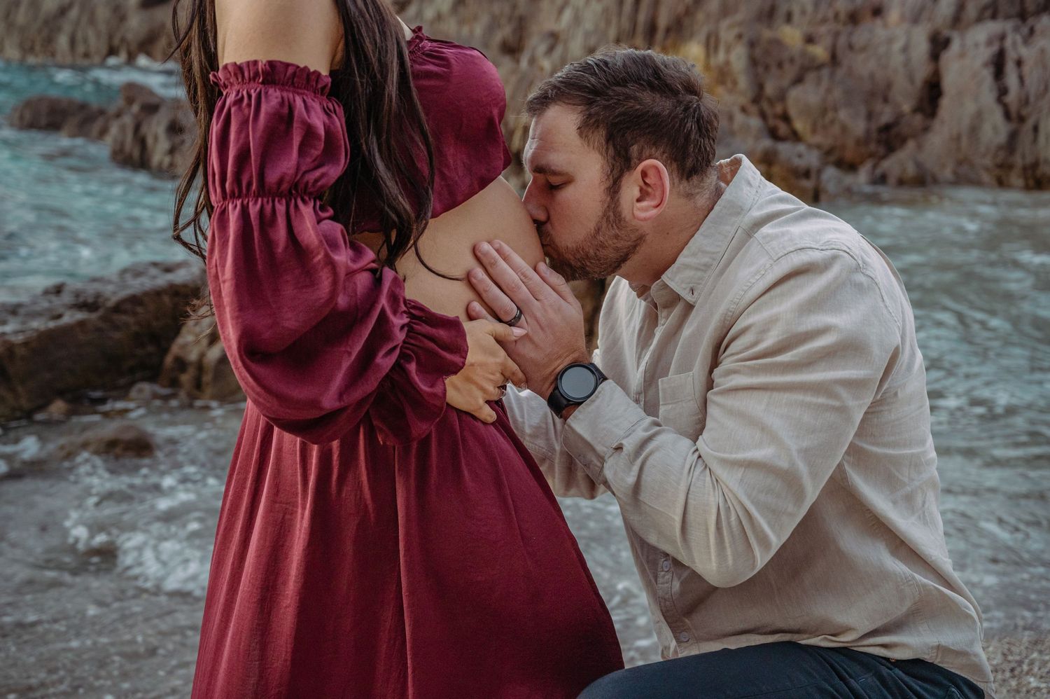 A tender moment during a maternity photoshoot as partner kisses the pregnant belly in a burgundy dress outdoors.