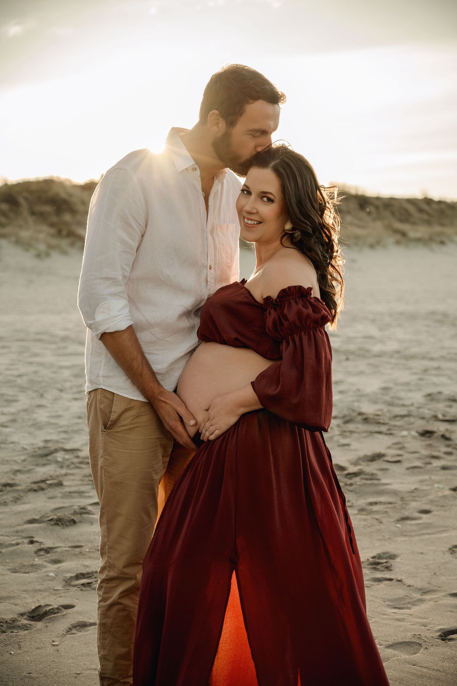 A romantic maternity photoshoot on the beach at sunset with a burgundy off-shoulder dress and soft natural lighting.