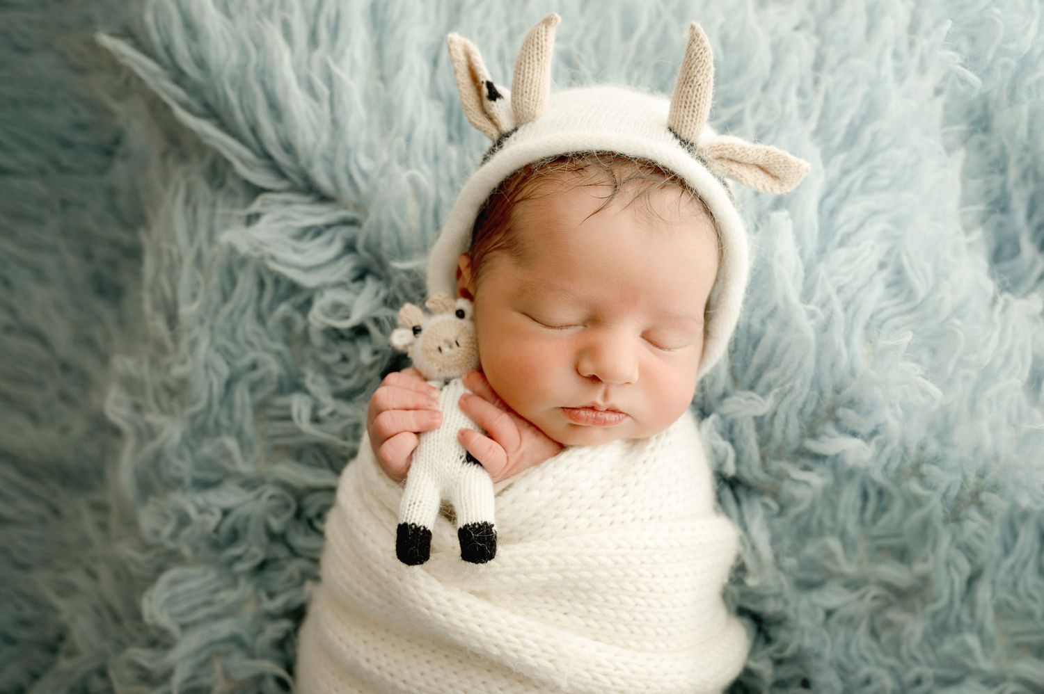 Sleeping newborn wrapped in cream-colored blanket wearing deer hat bonnet on fluffy blue backdrop.
