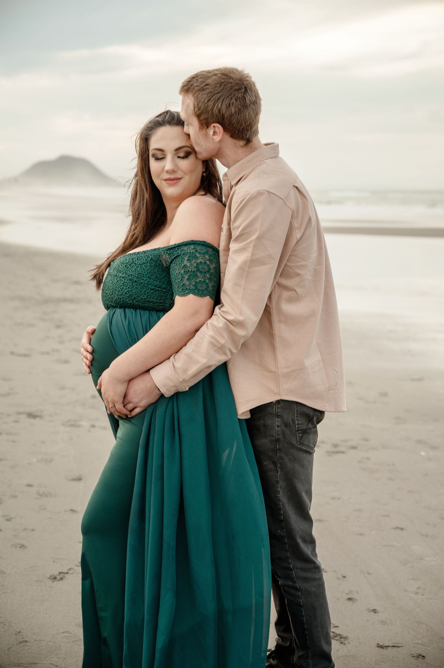 Expecting couple embracing on beach at sunset, woman wearing emerald green maternity dress.