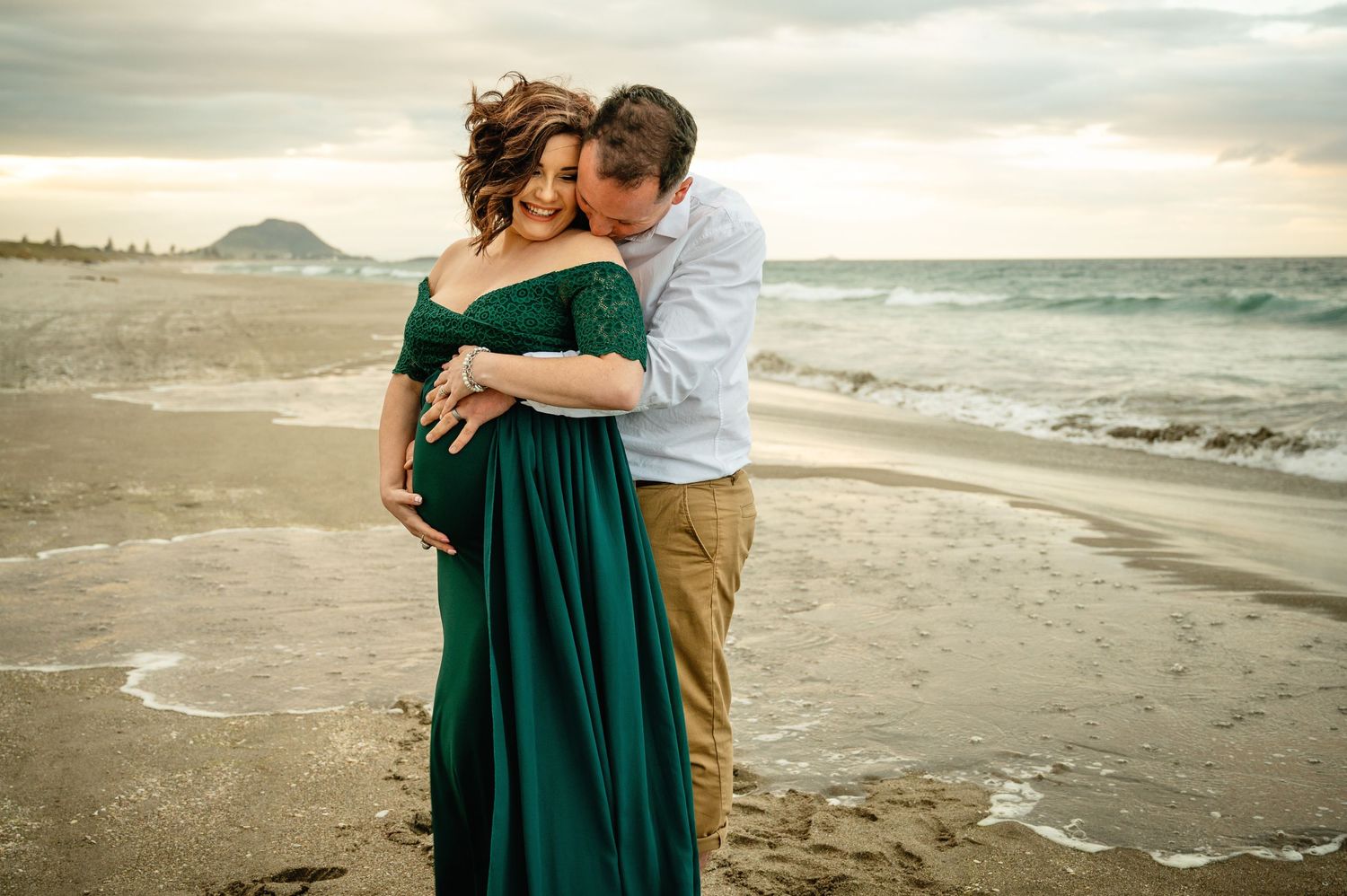 People embracing on a foggy beach during sunset with waves crashing in the background and mountains in the distance.