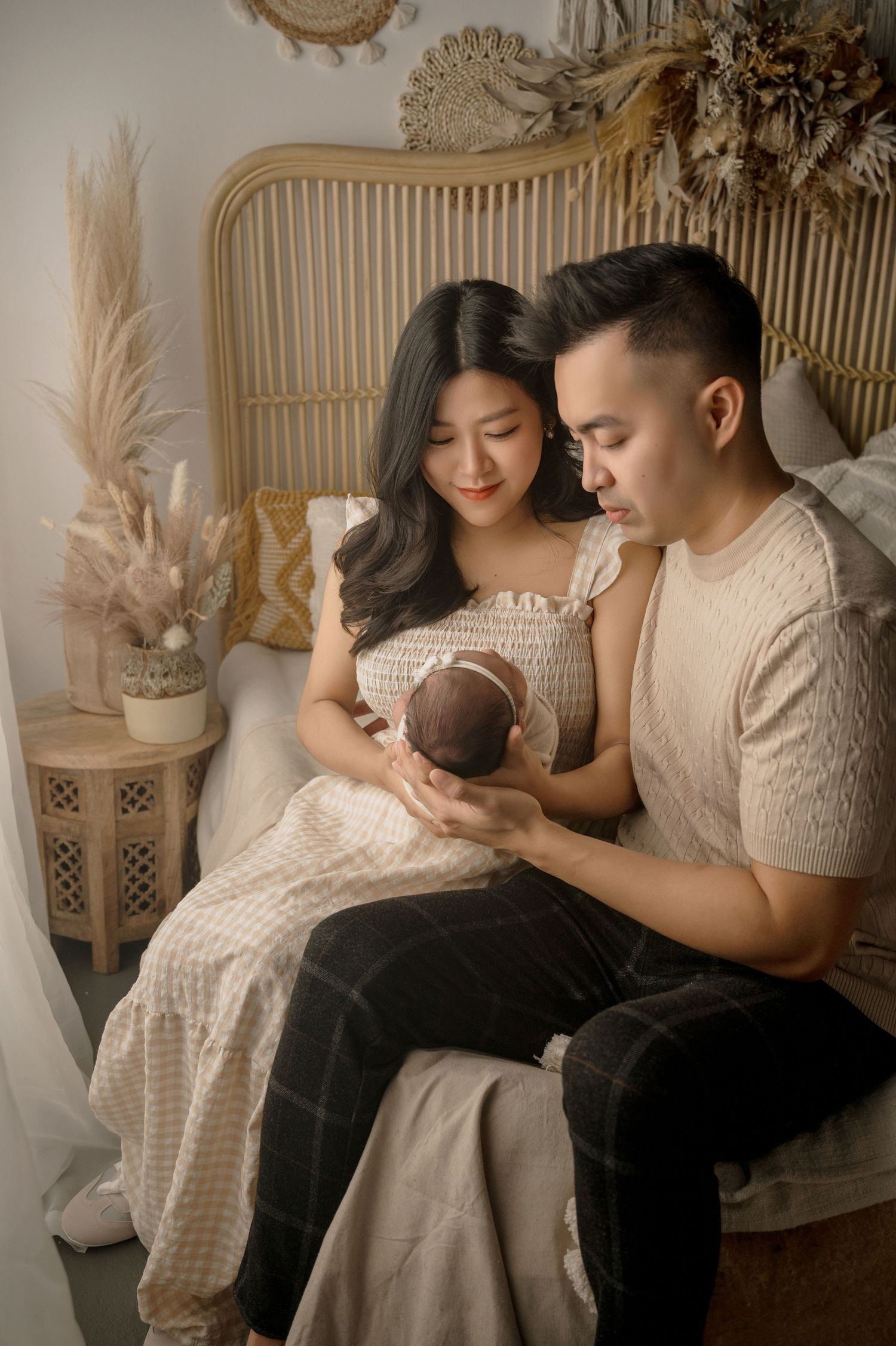 A couple in neutral toned sweaters sits together on a rattan chair with their newborn baby in a cozy home setting.