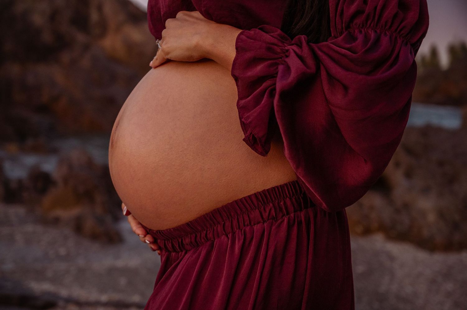 Close-up of a pregnant belly in a flowing burgundy dress against a natural backdrop.