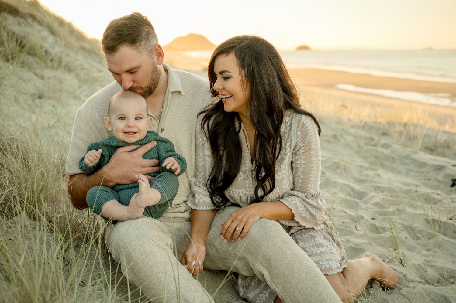 Family sits together on grassy beach dunes during golden hour sunset photo session.