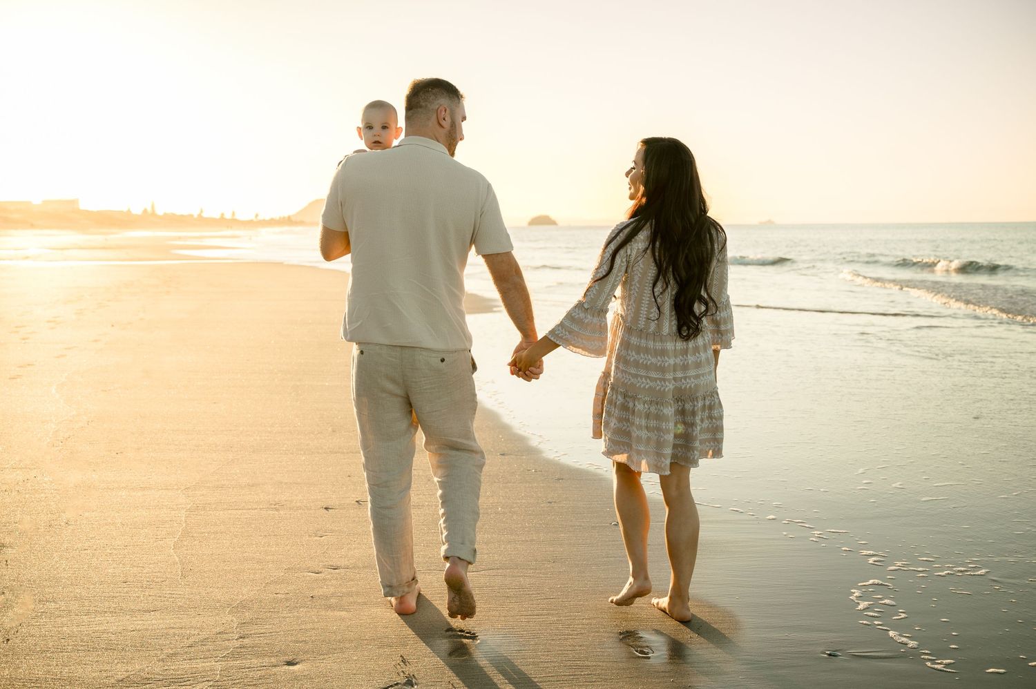 A couple walks hand in hand along a sandy beach at sunset with golden light illuminating their path.