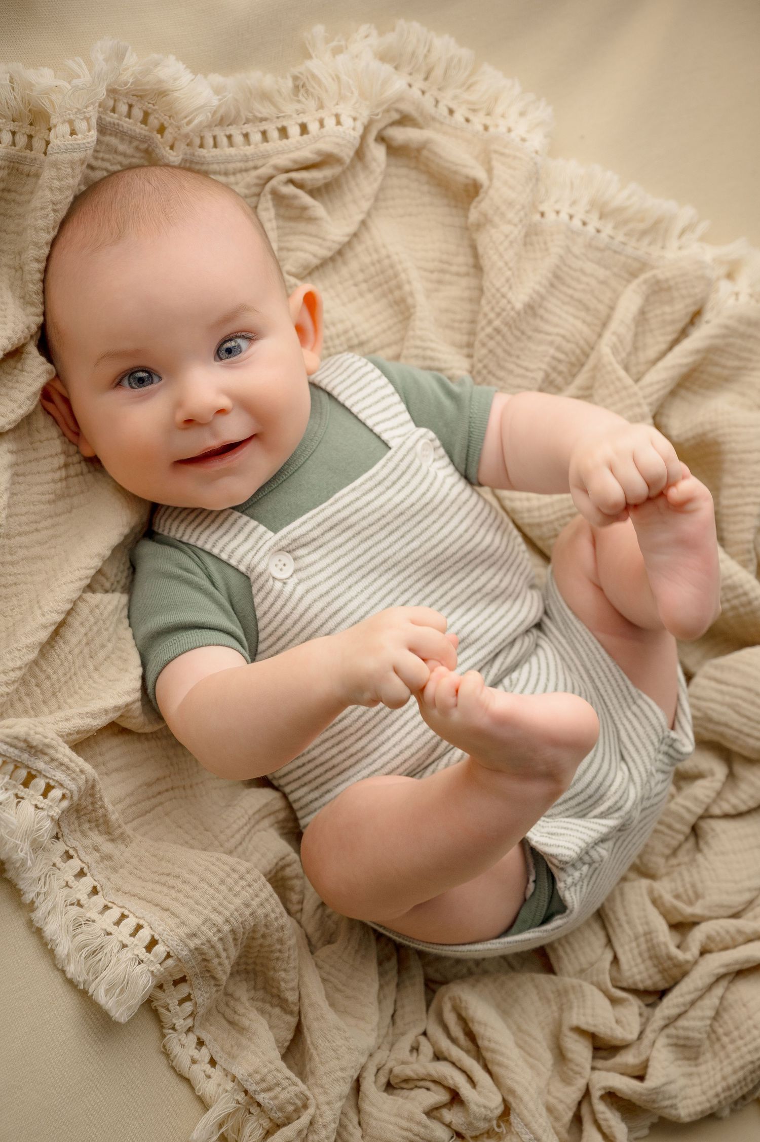 A baby in green and striped overalls lies on cream textured blanket during professional photoshoot.