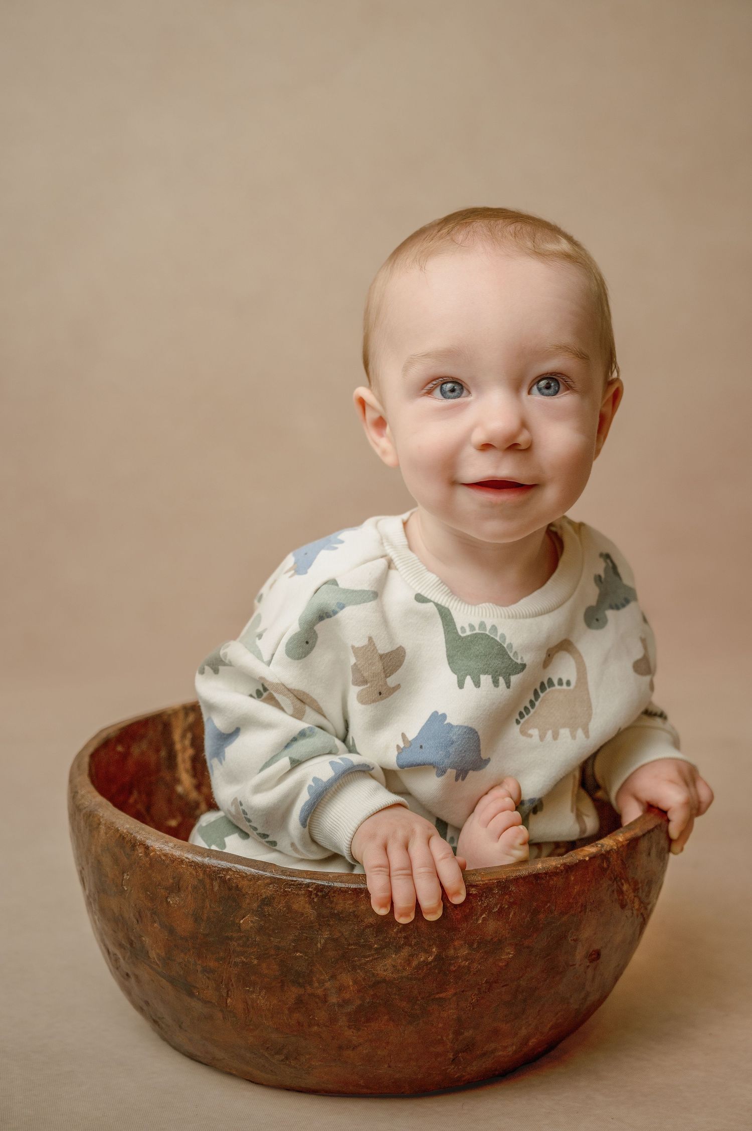 Baby wearing dinosaur print onesie sits in wooden bowl against neutral backdrop.