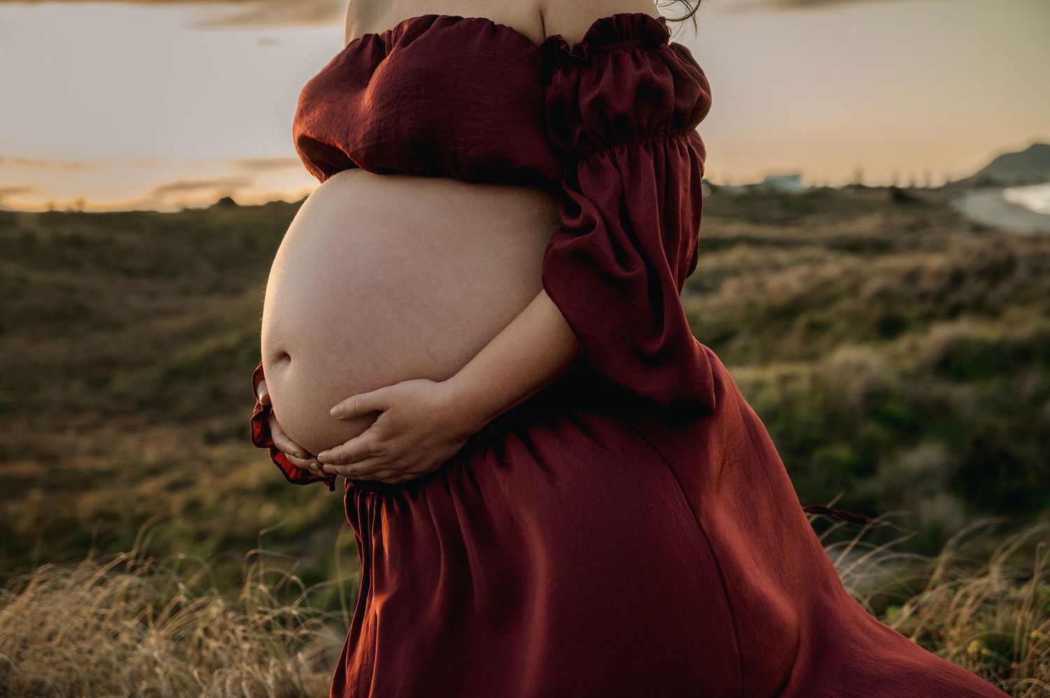 Close-up of a pregnant belly draped in a burgundy dress against a natural outdoor backdrop.