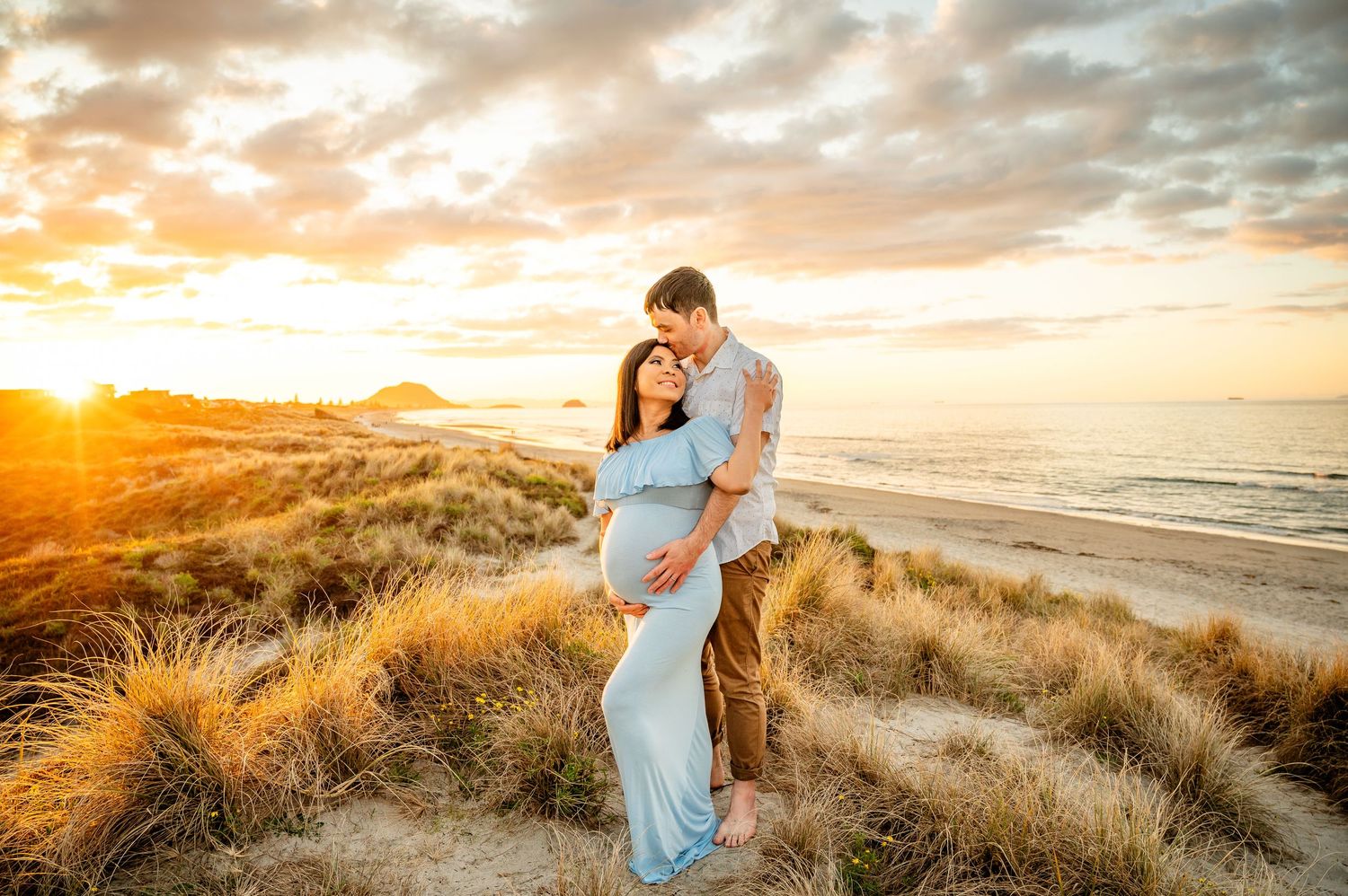 Couple stands against stunning golden sunset at beach with tall grass dunes creating romantic coastal atmosphere.