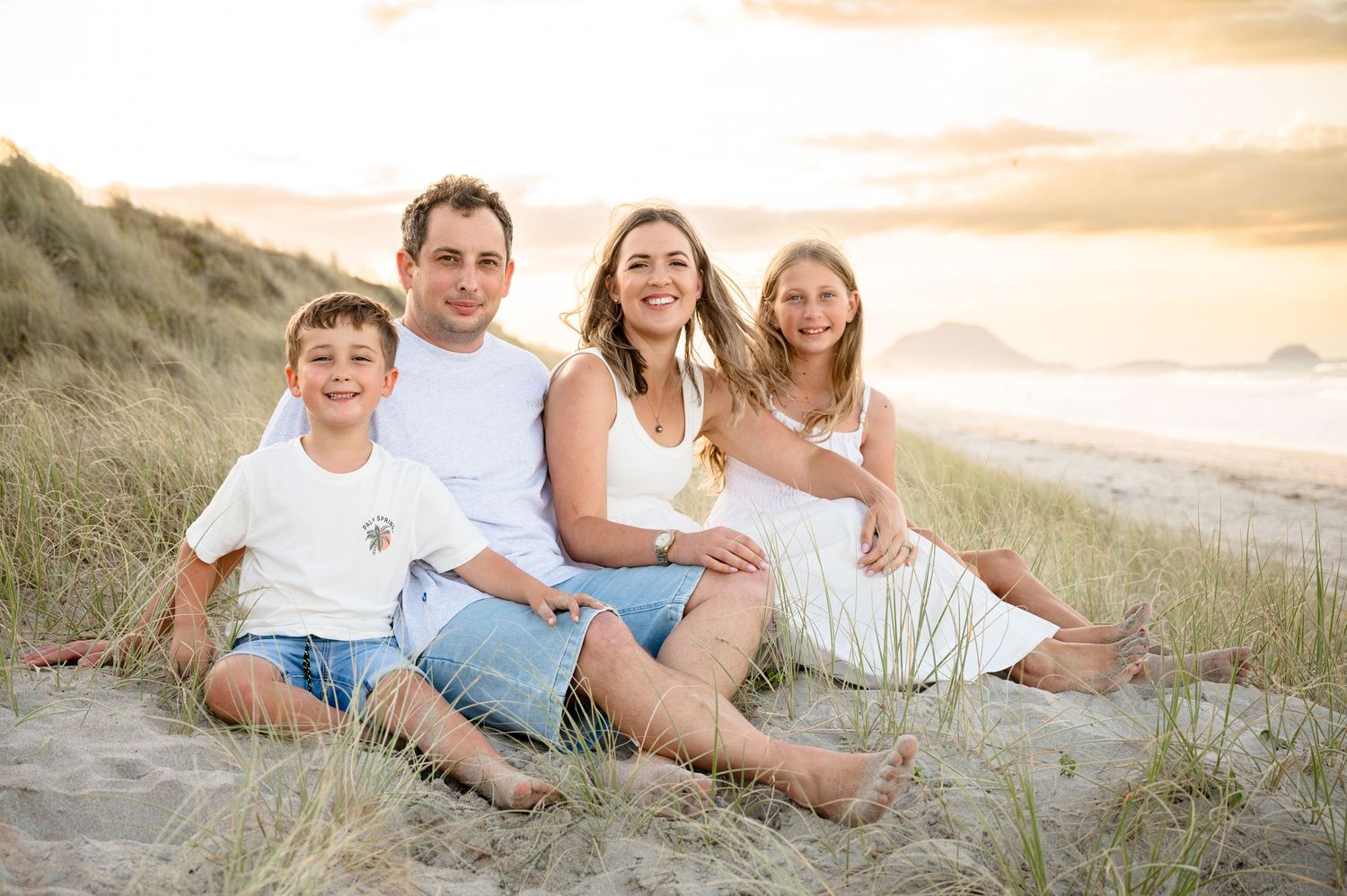 A family sits together on sandy dunes at sunset with ocean waves visible in the background.