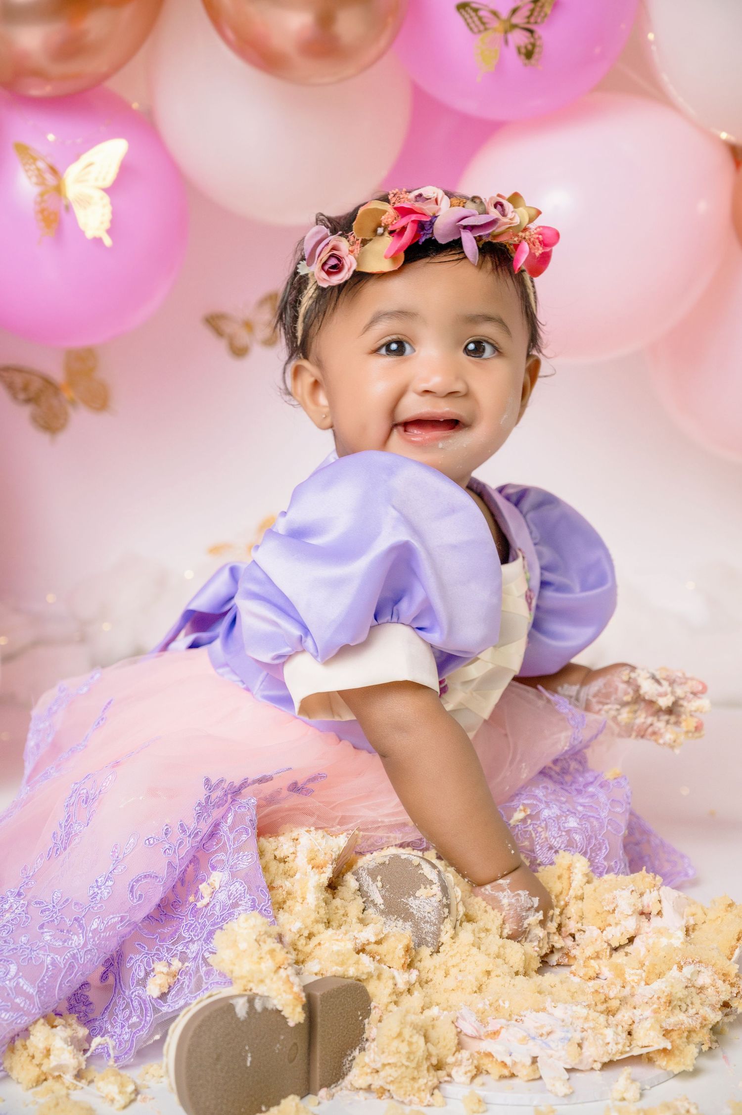 Baby in purple dress and floral crown smiling among pink balloons with gold butterflies during cake smash session.