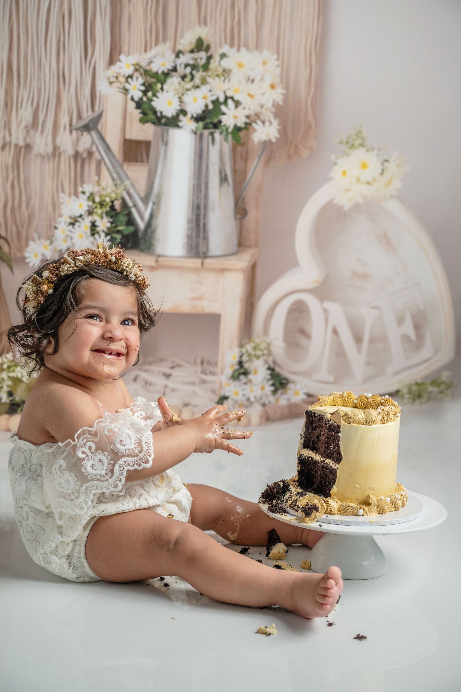 A baby in white lace outfit sits next to birthday cake with floral decorations and watering can.