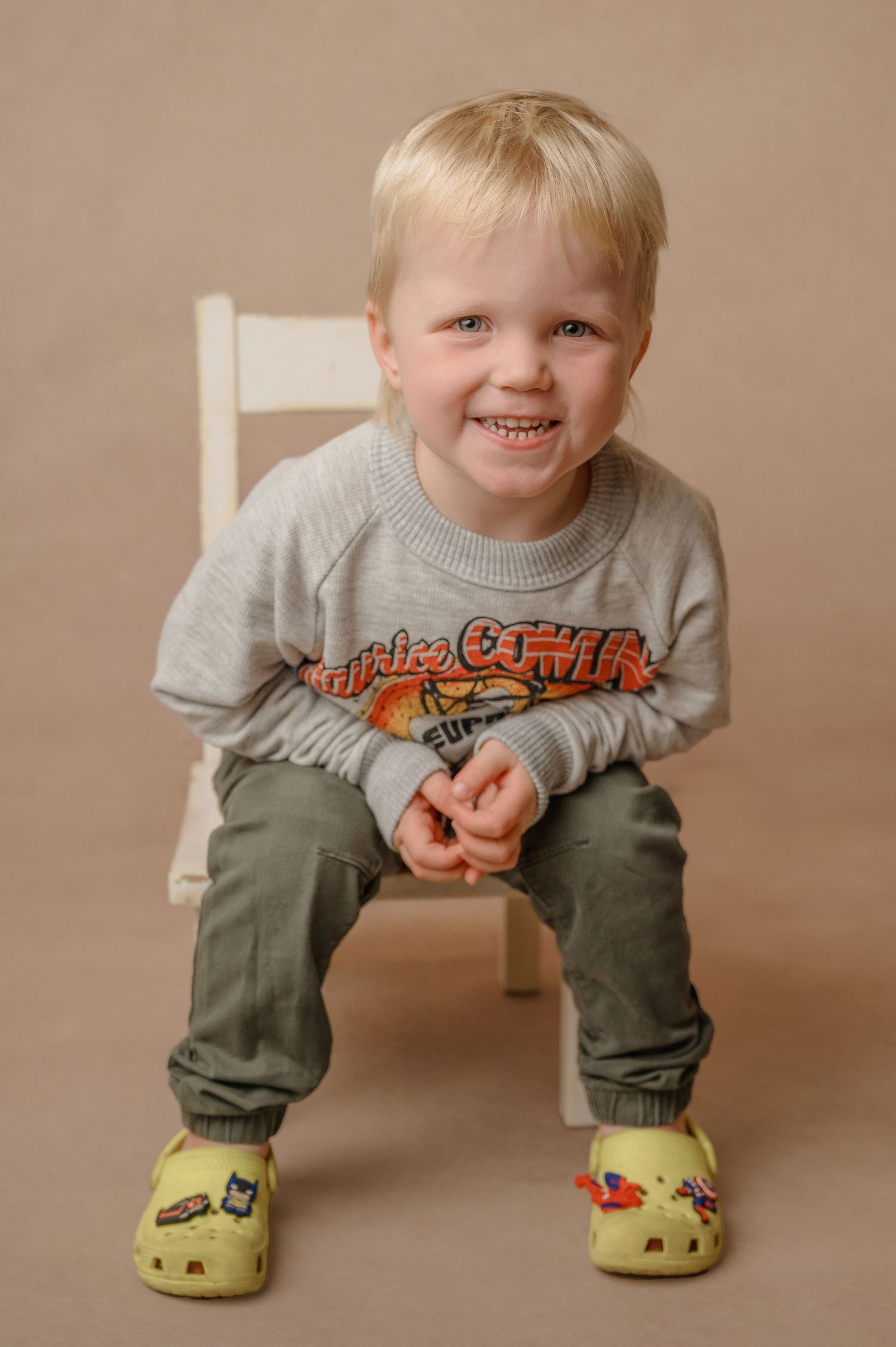 Young child wearing a gray sweatshirt and green pants sits smiling on a white chair wearing yellow crocs.
