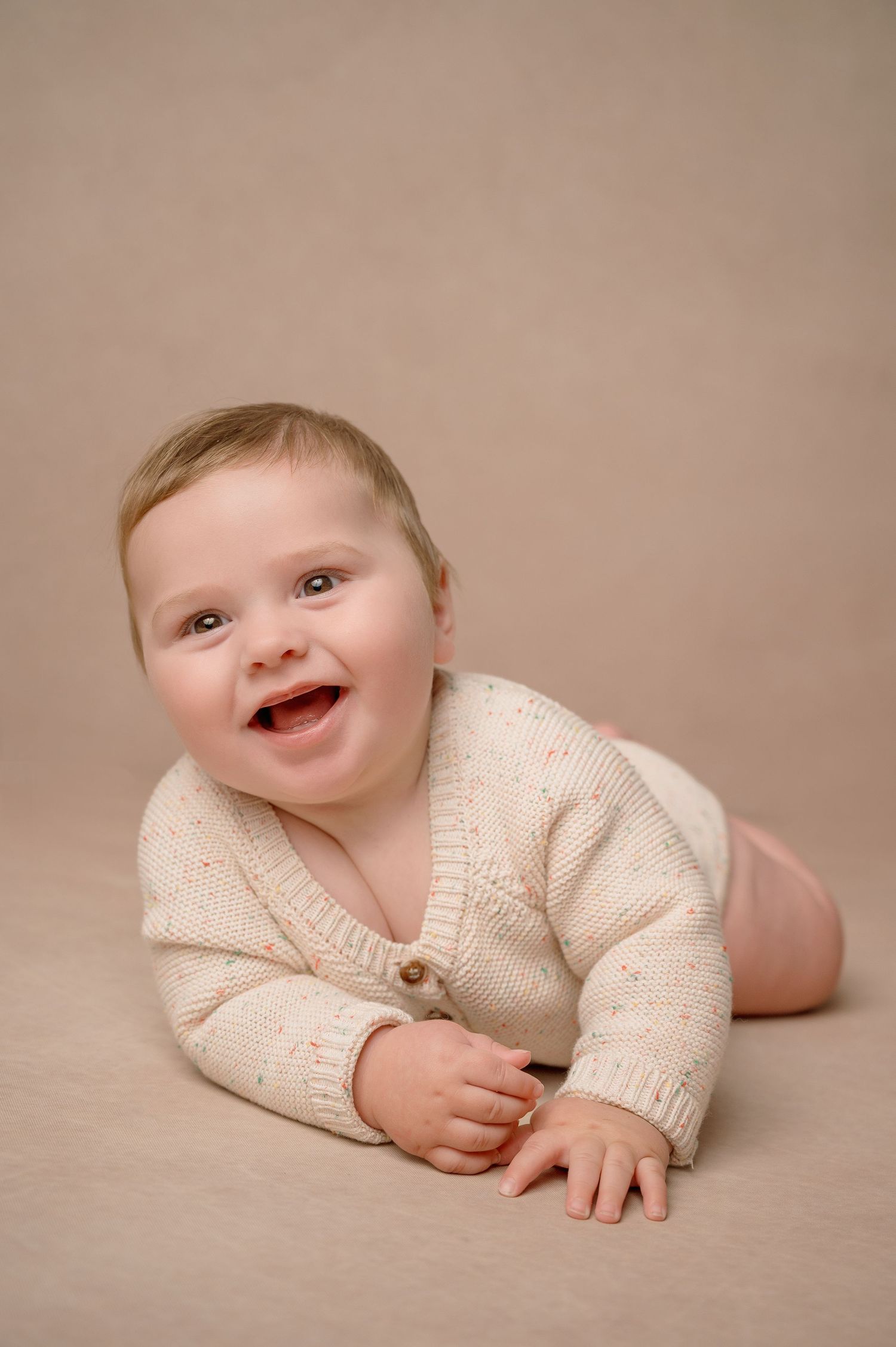 A baby wearing a cream knit cardigan lies on a beige background during a portrait session.