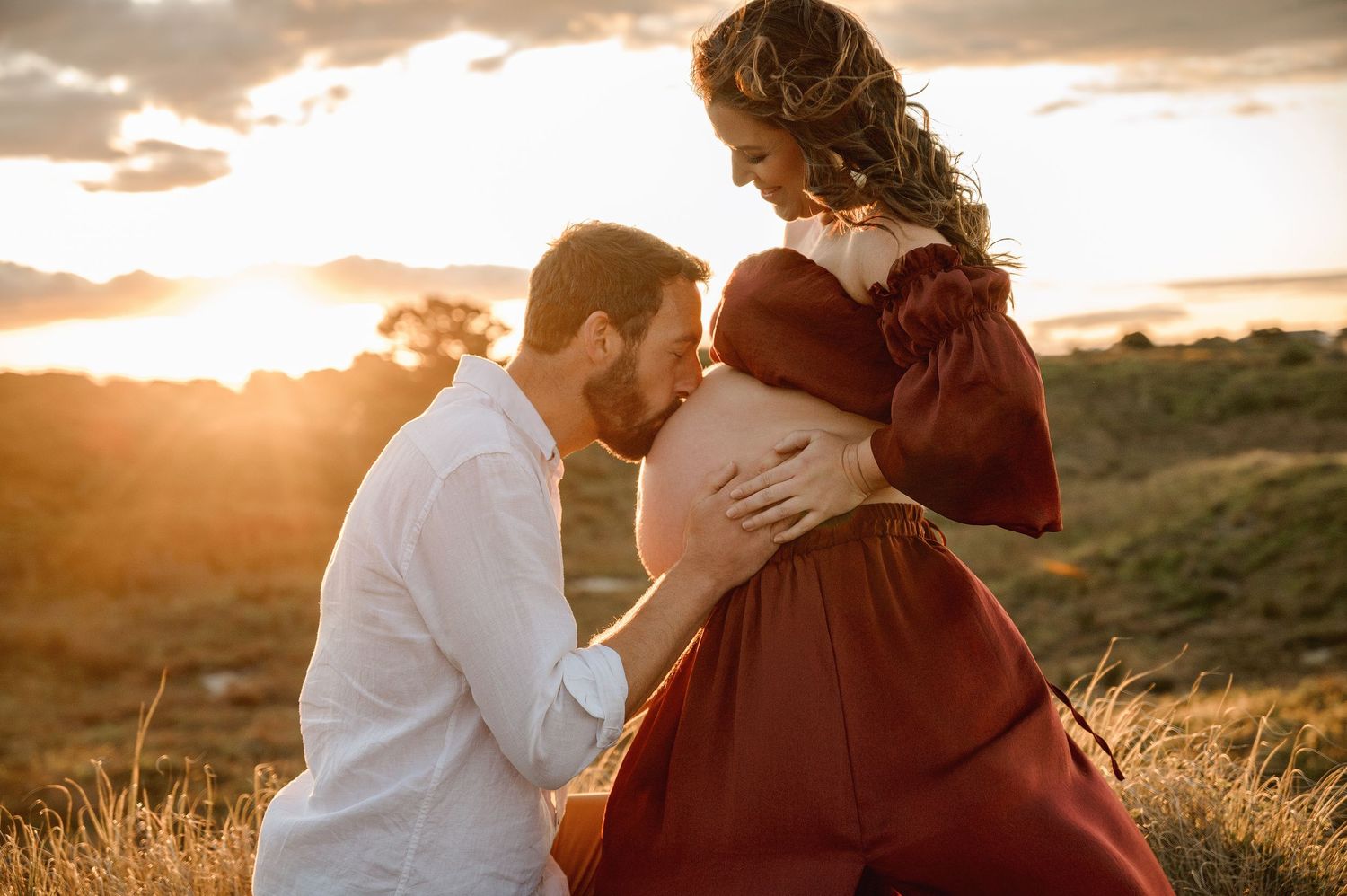 Sunset maternity photo with partner kissing baby bump in burgundy dress on grassy hillside.