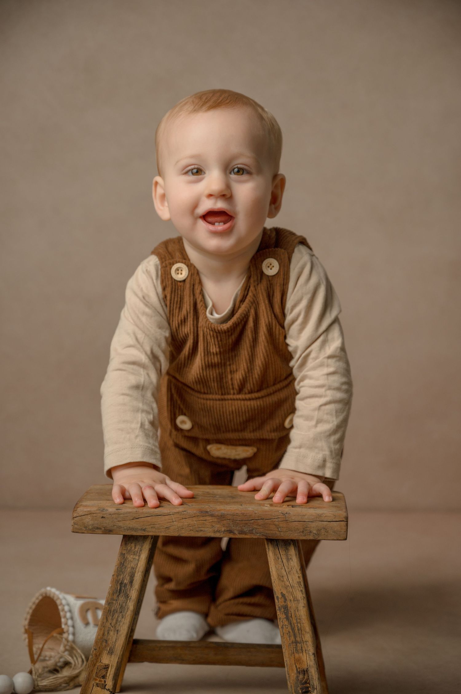 A happy toddler wearing brown corduroy overalls stands at a rustic wooden stool during a studio portrait session.