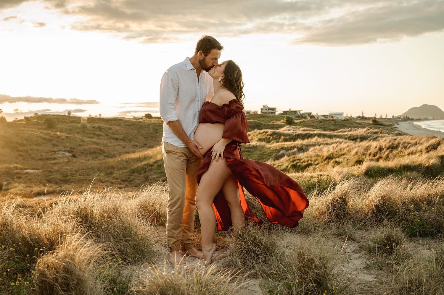 Romantic couple sitting in coastal grass dunes during golden hour with ocean views in the background.