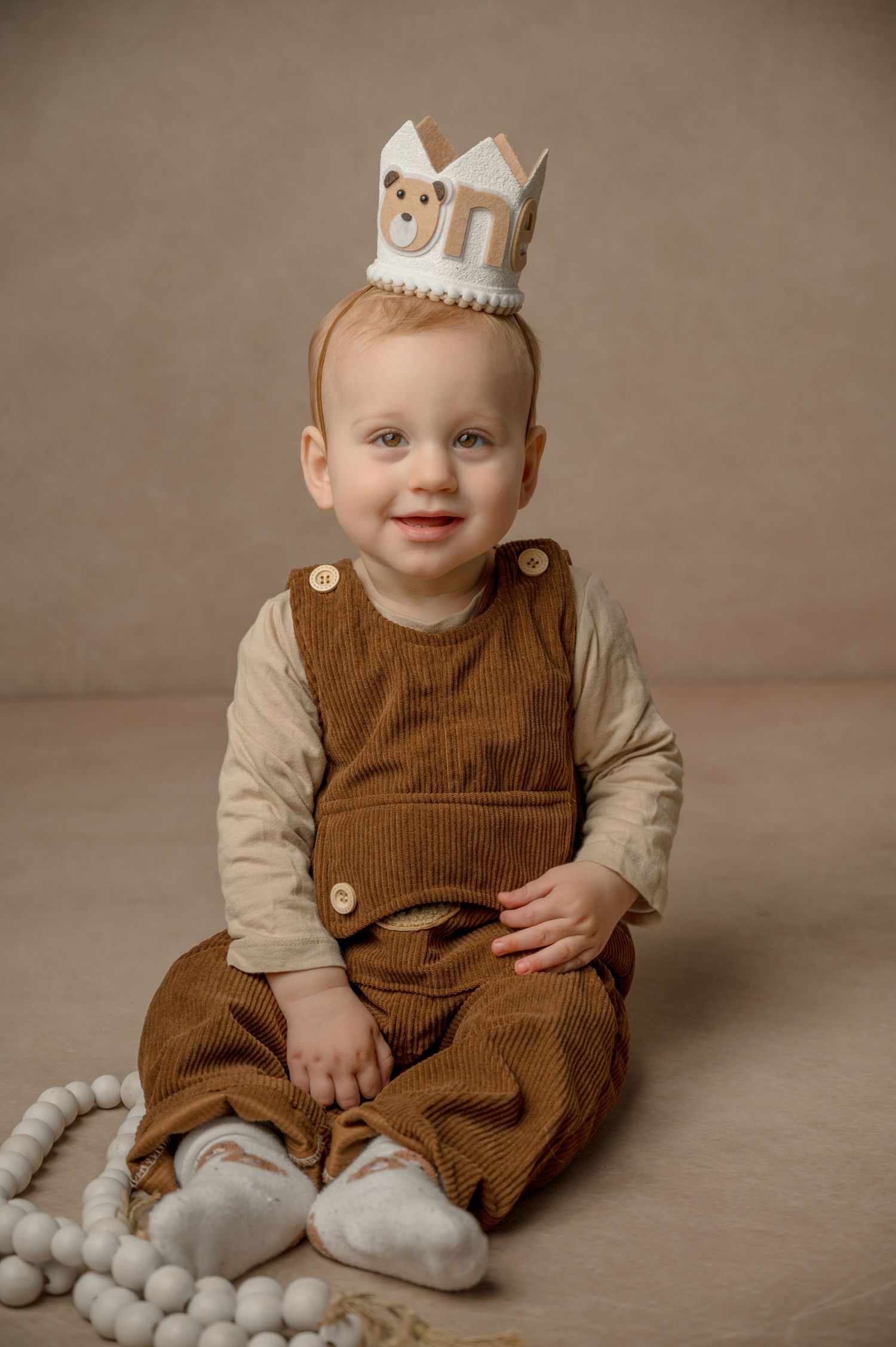 Baby wearing brown corduroy overalls and beige shirt sits on floor wearing decorative paper crown in studio portrait.
