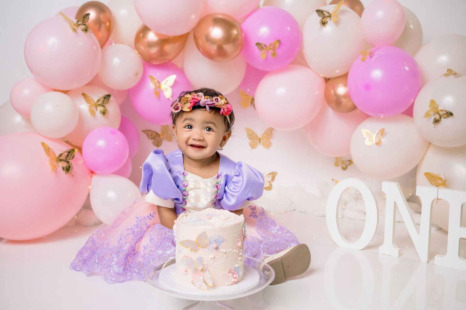 A birthday setup with pink and gold balloon arch, butterflies, and a cake smash scene against a light pink backdrop.