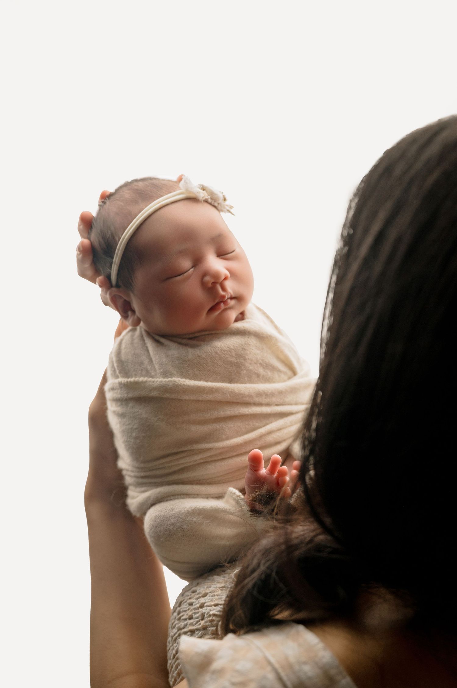 A newborn baby wrapped in a soft beige swaddle blanket sleeps peacefully while being held against a light background.