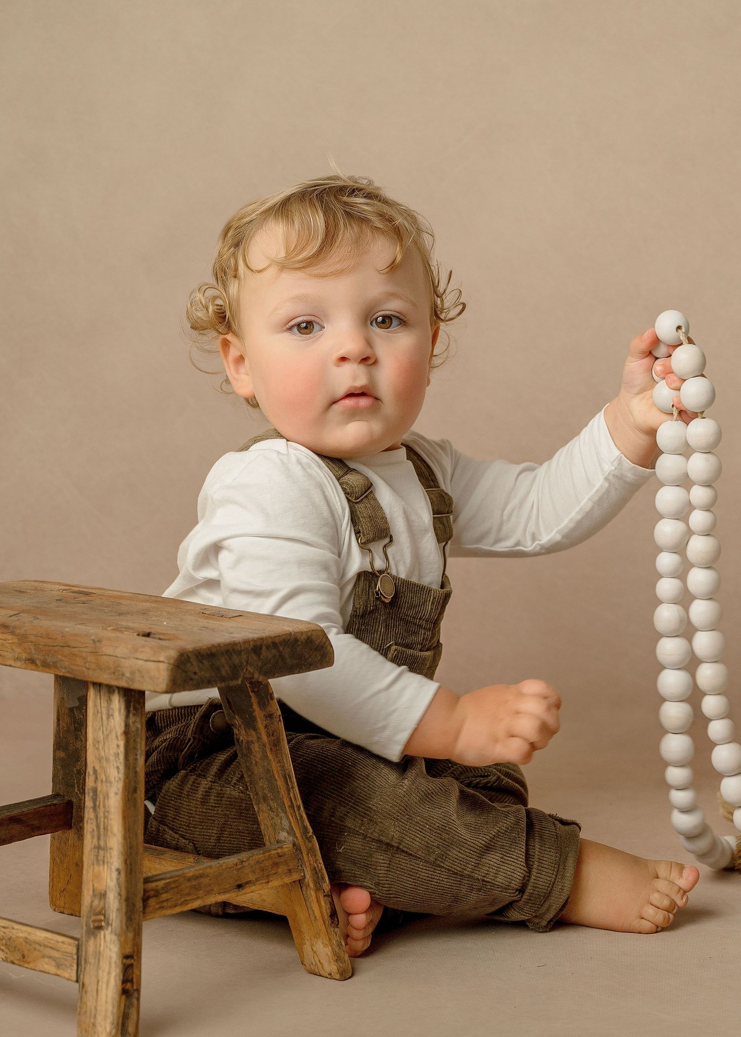 Toddler in overalls sits on wooden stool holding white beaded necklace against beige background.