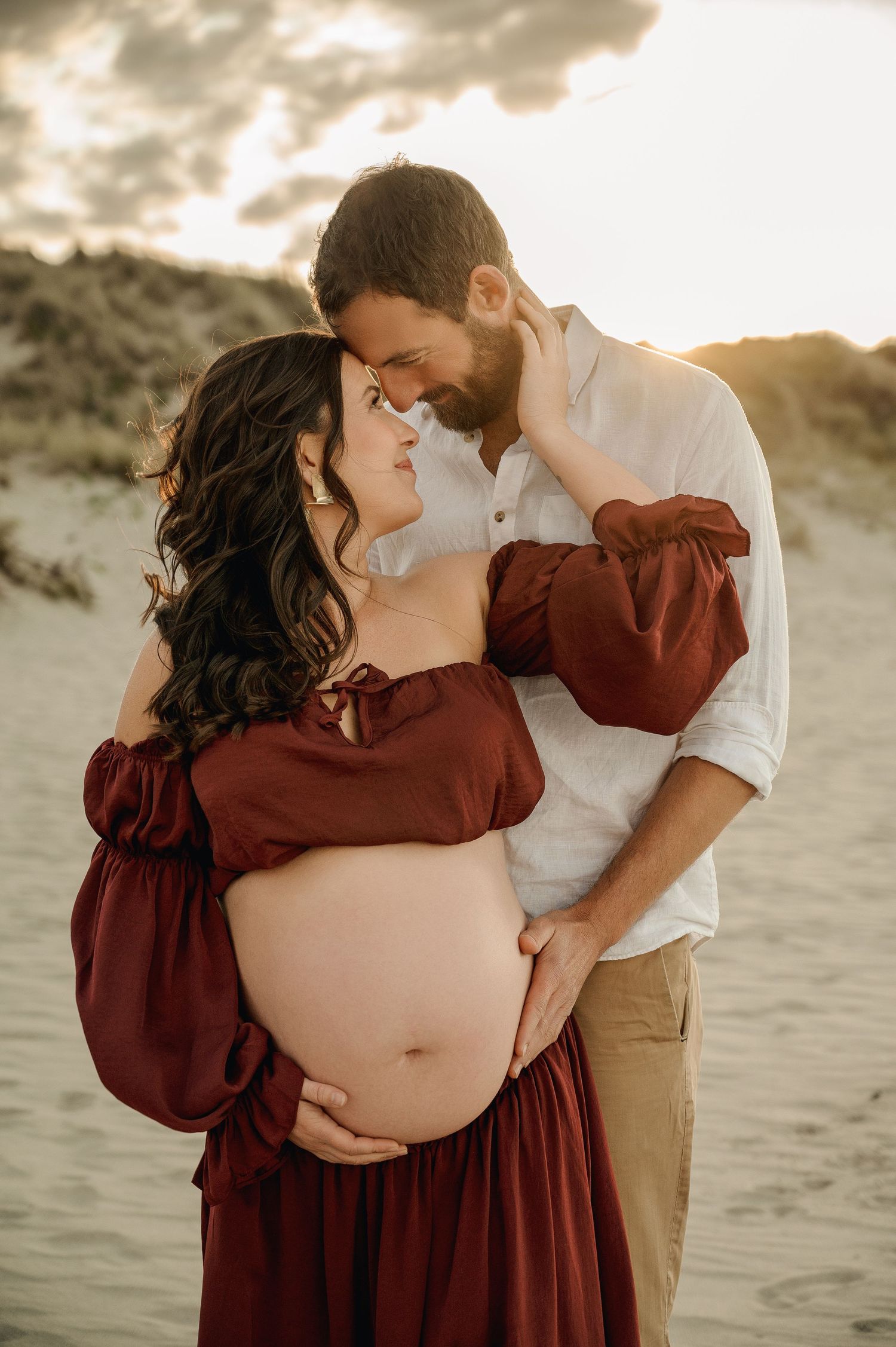 A romantic maternity photo session on the beach at sunset features a couple in burgundy dress and white shirt embracing tenderly.