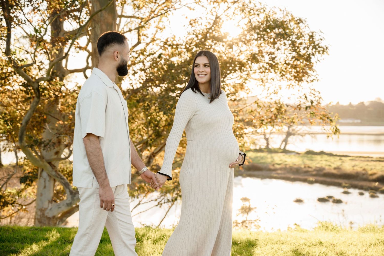 A couple in matching white outfits holds hands by a lakeside during golden hour sunset in a romantic outdoor setting.