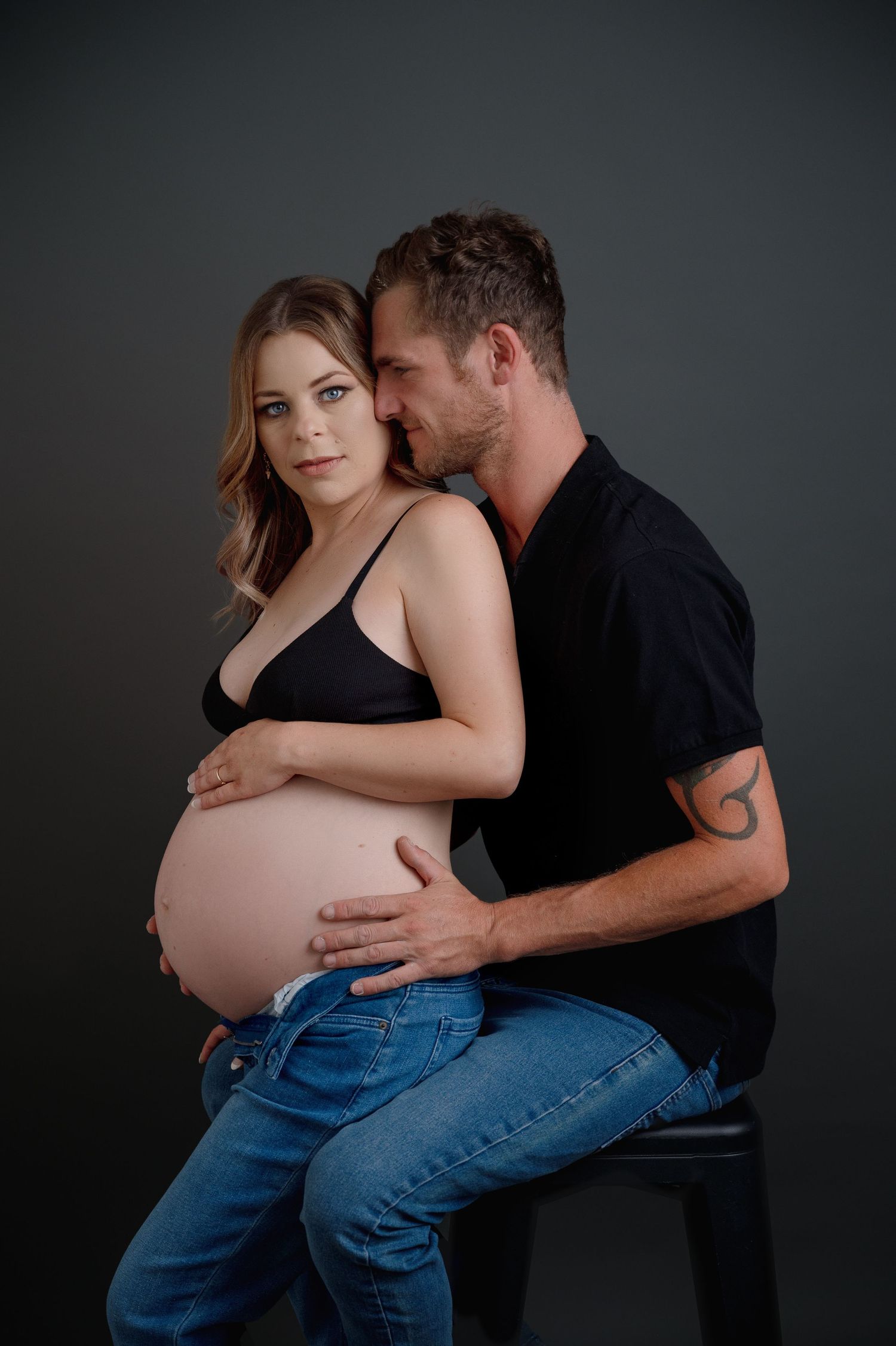 Maternity photo series showing couple embracing against dark background in casual black and denim attire.
