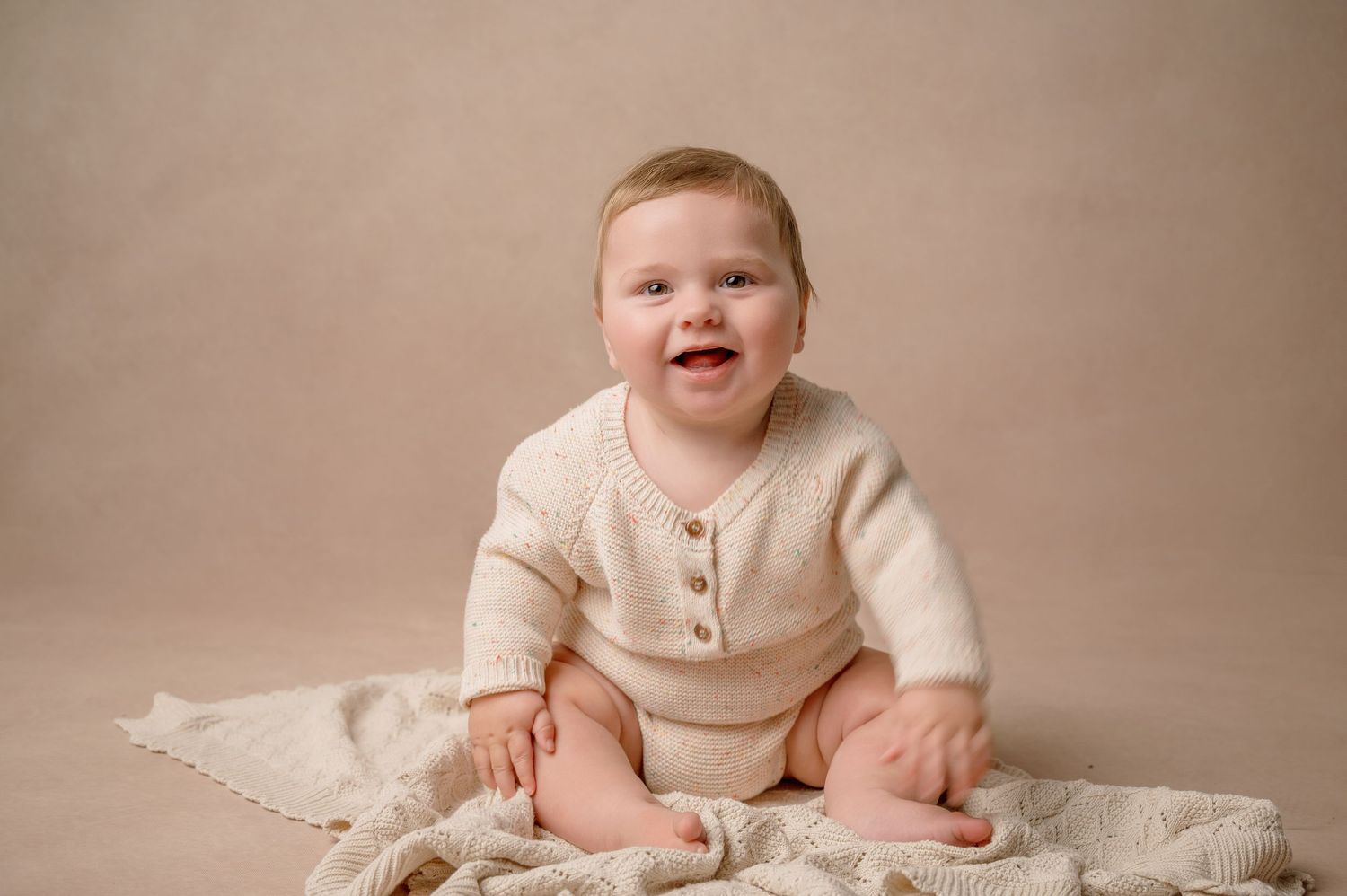 Baby wearing cream henley top sits on textured blanket against neutral beige backdrop.