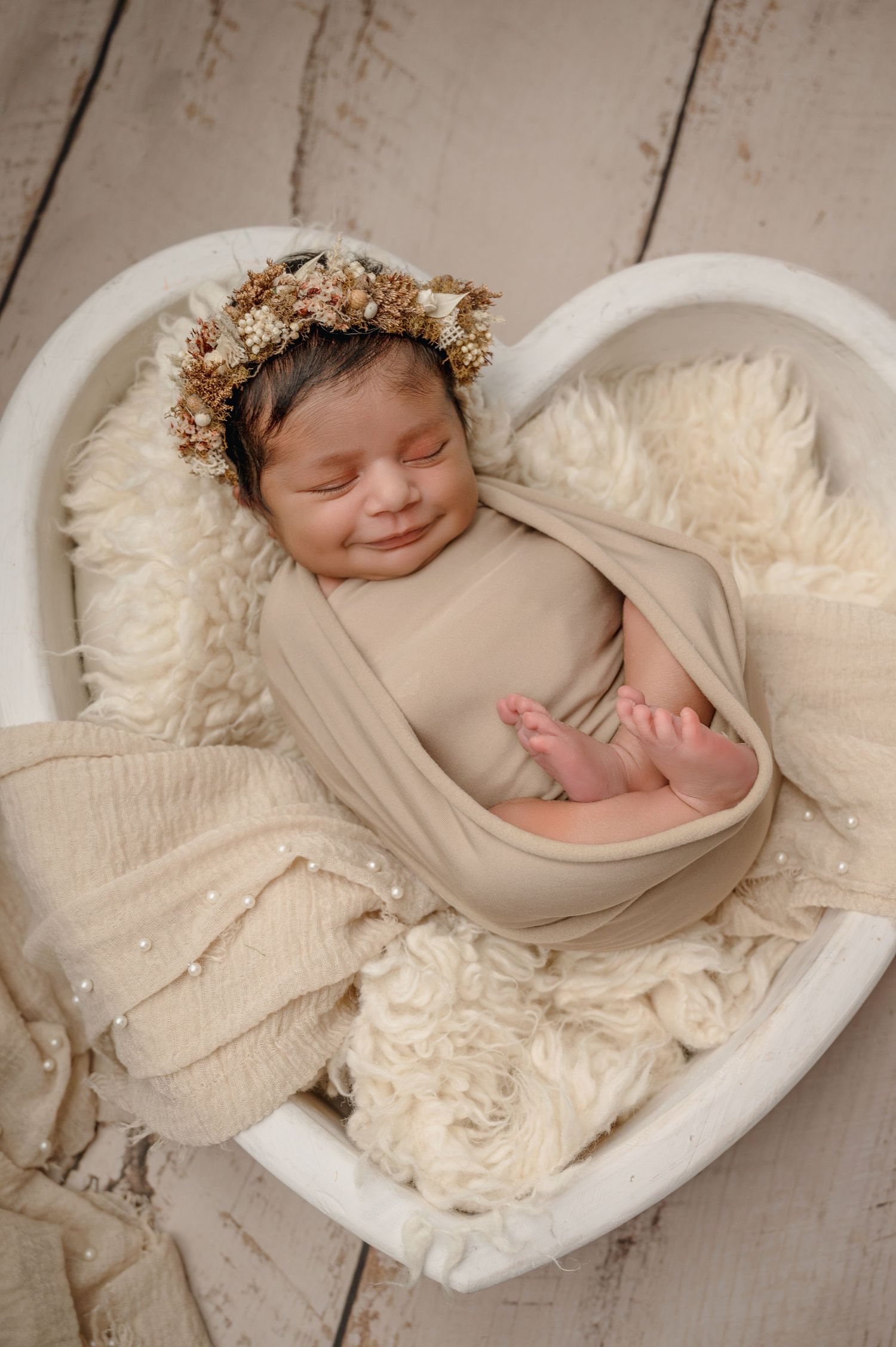 Sleeping newborn in beige wrap posed in heart-shaped bowl with floral crown and textured backdrop.