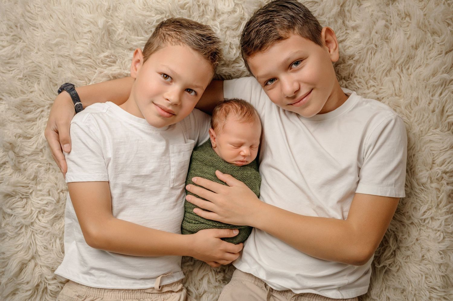 Family members wearing white t-shirts lie together in warm embrace against textured beige backdrop.
