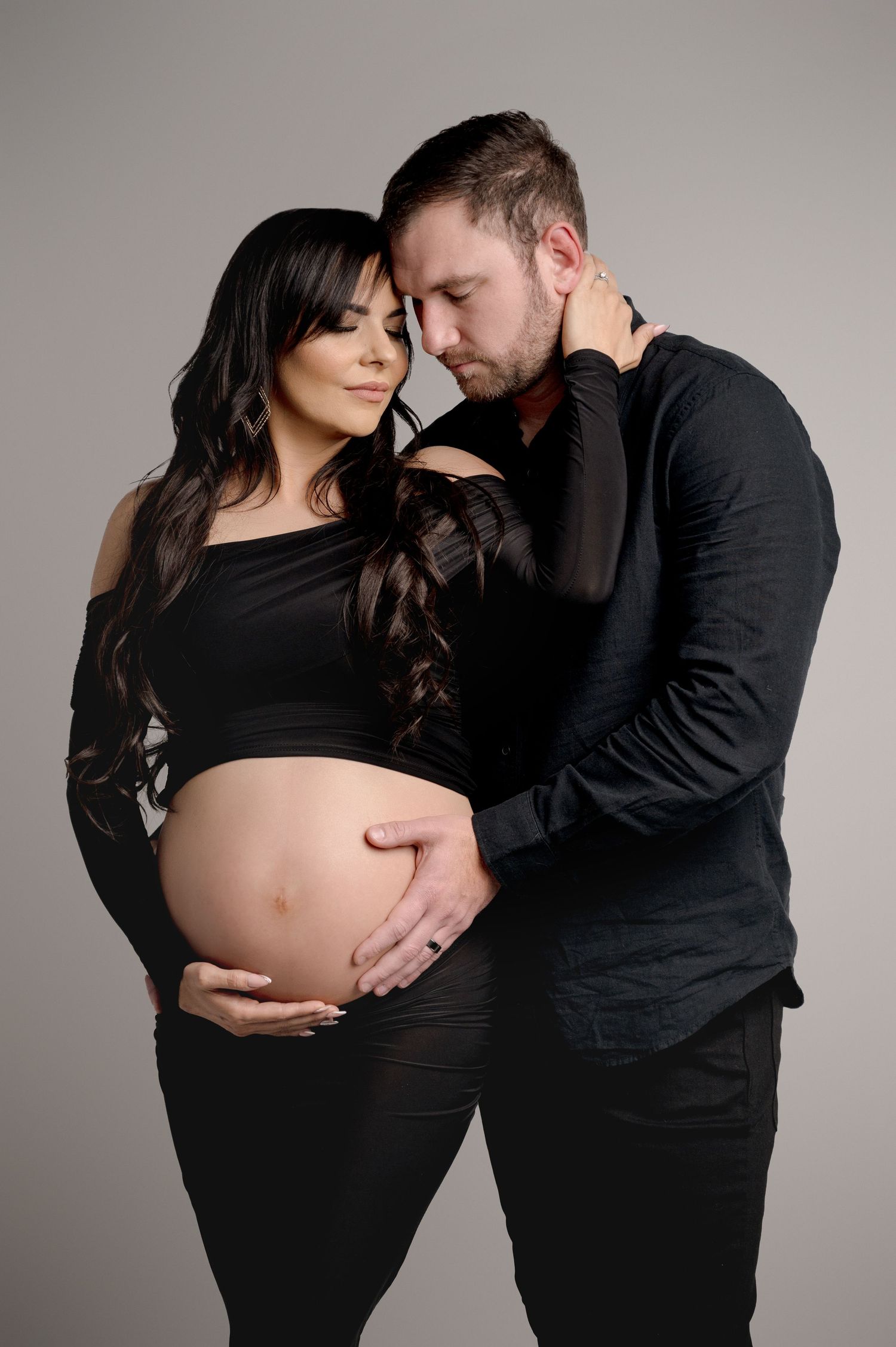 Intimate maternity photo of couple in black outfits against grey studio backdrop.