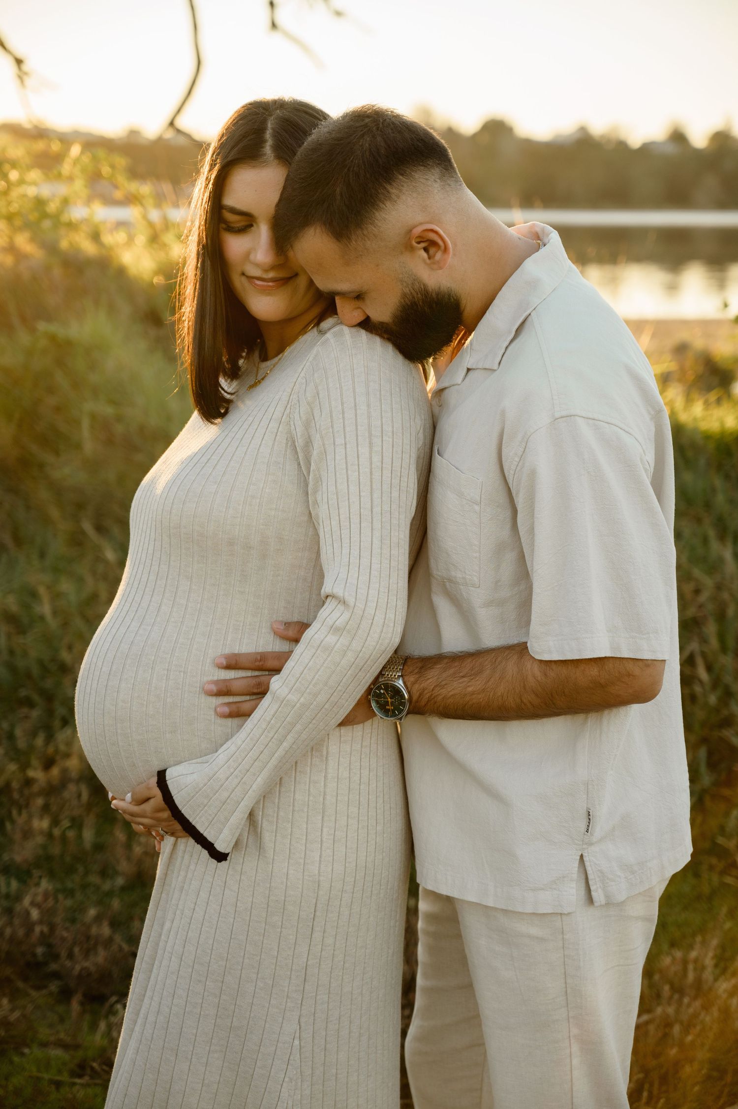 Expectant couple shares intimate moment outdoors at golden hour in matching white outfits during maternity photoshoot.