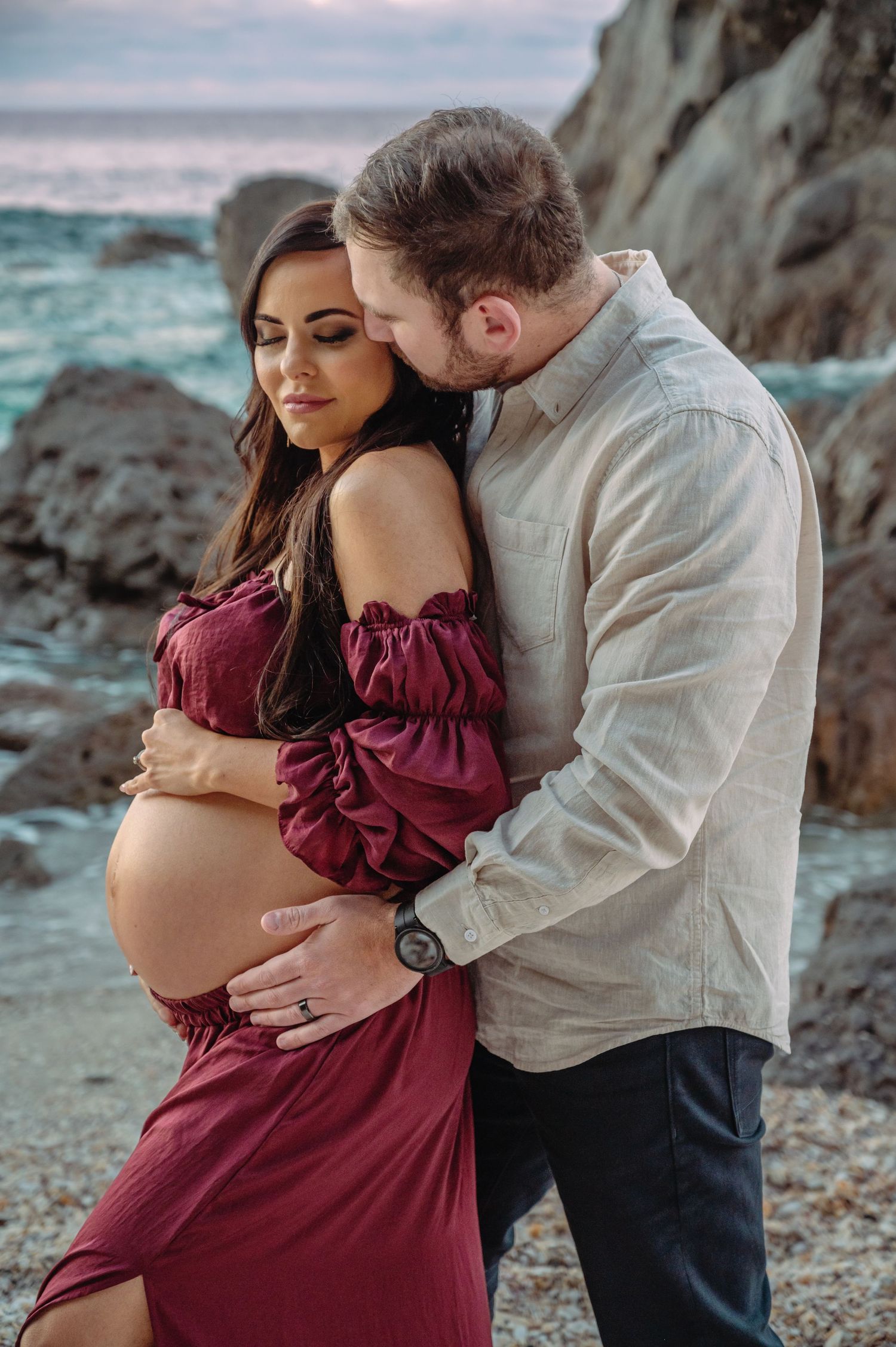 Maternity photo of couple by rocky coastal shore, woman in burgundy dress at sunset.